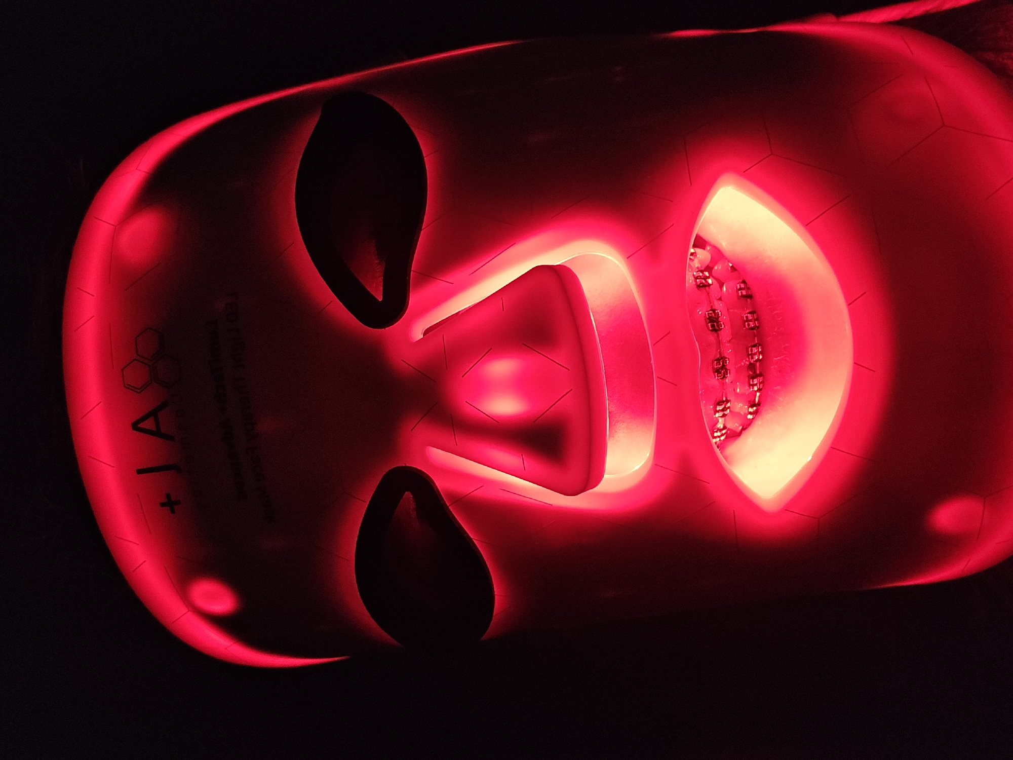 A person is depicted wearing a red light therapy face mask in a dark setting. The mask, covering the entirety of the face with cutouts for eyes, nose, and mouth, is illuminated intensely from within by numerous red LED lights. This creates a strong red glow that highlights the contours of the mask and the features of the wearer's face contained within.
A notable detail is the presence of dental braces clearly visible on the wearer's teeth, reflecting the red light emanating from the mask's mouth opening. The image is captured from a slightly elevated perspective and rotated 90 degrees clockwise. The background is uniformly black, enhancing the visual impact of the bright red mask.
On the mask's right cheek area (visible on the left side of the image due to rotation), partial text and a logo are discernible. The text includes "+JA" or similar characters, followed by a logo resembling interconnected hexagons. Below this, smaller, less legible text, also rotated, appears to be product branding, possibly including phrases like "For All" and "LED MASK." The mask itself has a smooth surface with faint geometric patterns. The overall scene suggests a personal wellness or skincare treatment session utilizing advanced light therapy technology.