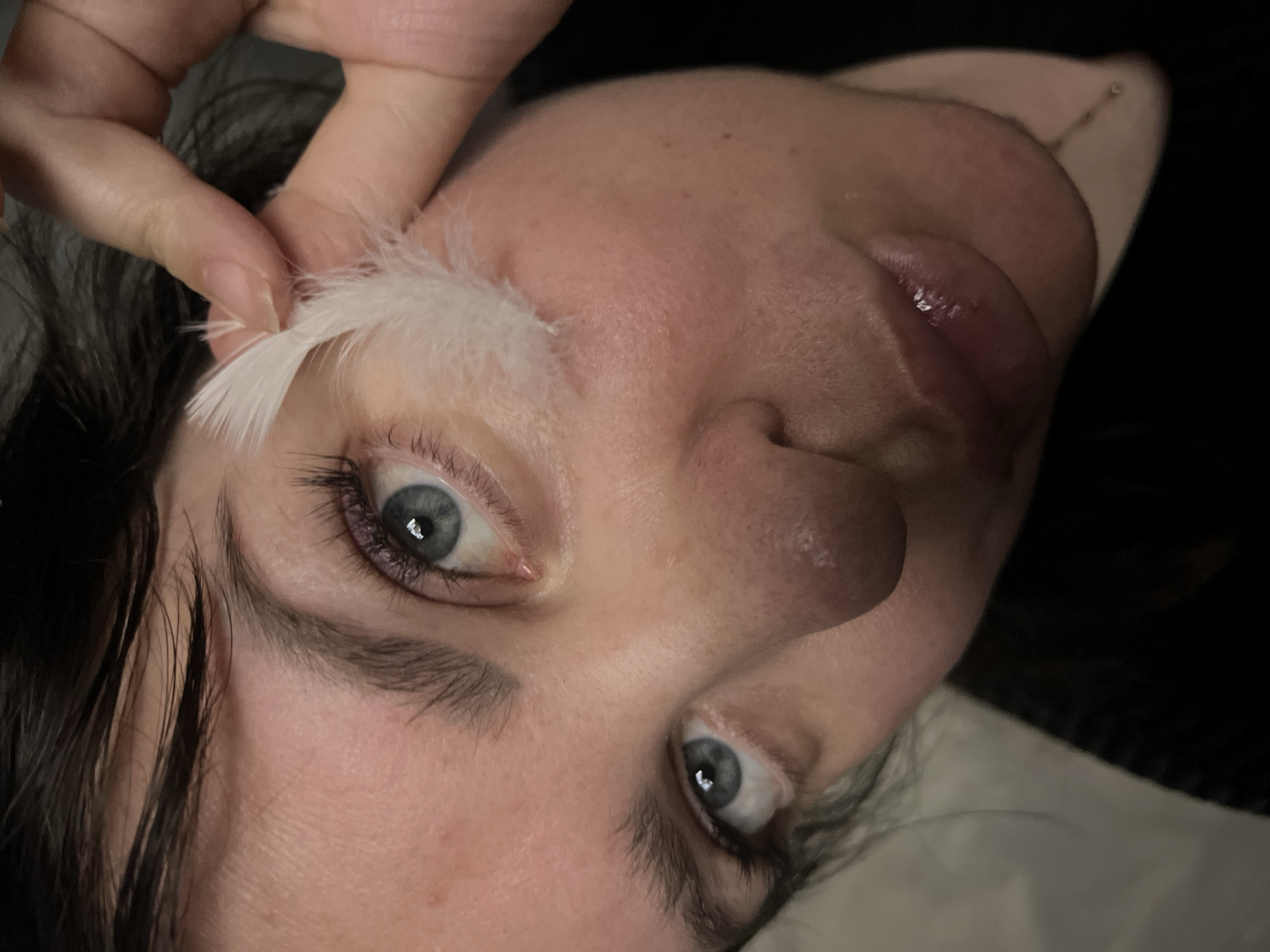 A close-up, low-angle shot captures a person's face, focusing on their eyes and lips. A small white feather is held near the person's right eye, with the index finger and thumb of their right hand. The person's dark hair is visible along the top edge of the frame. The background is dark and out of focus. There is no information about the location in the city of Noviy, Russia.