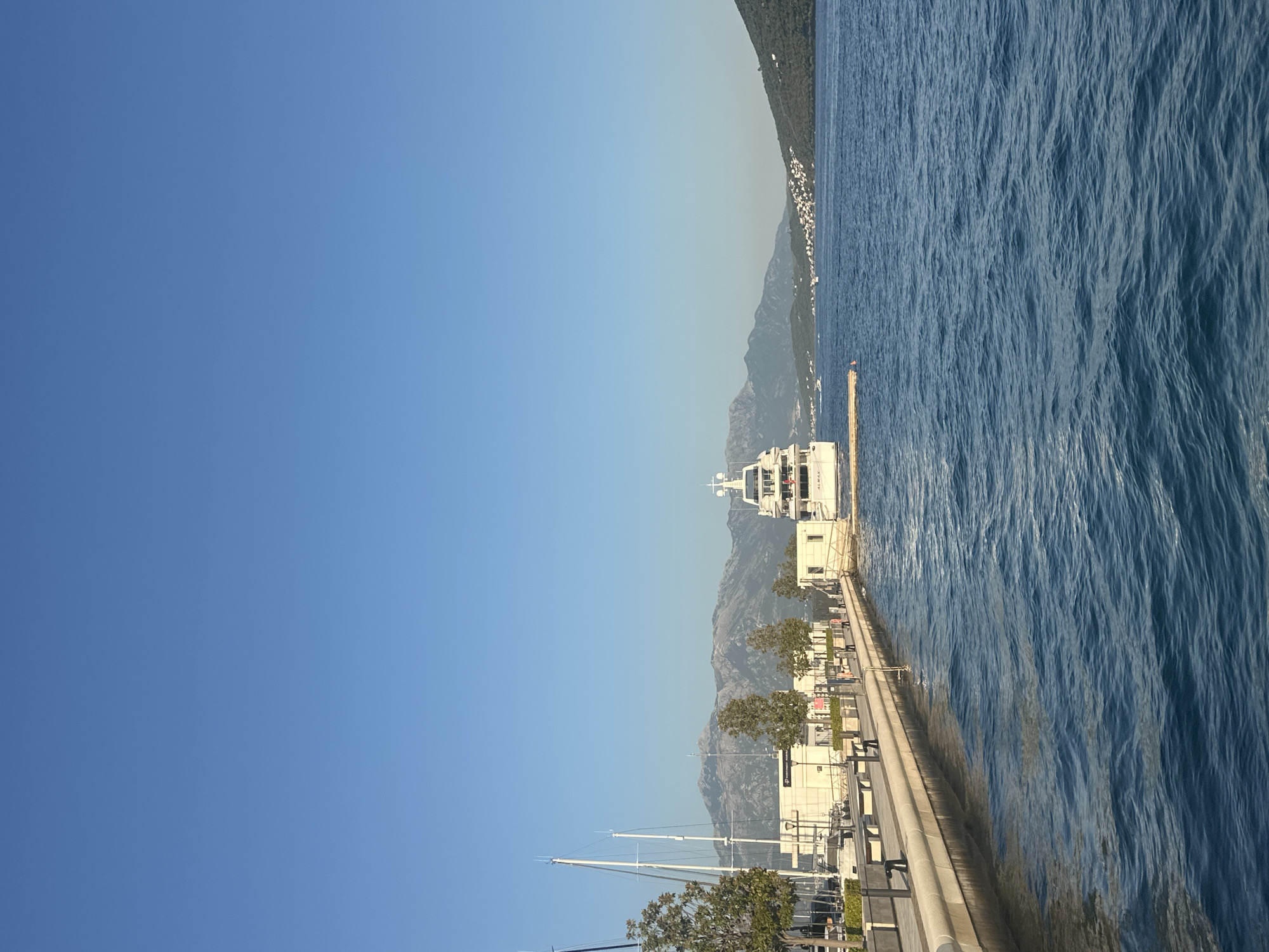 The image depicts a harbor scene in Herceg Novi, Montenegro. The scene is dominated by a clear blue sky and calm, deep blue water. A modern white yacht is docked along a paved promenade, which features several white modular buildings, streetlights, and trees. A line of benches is visible along the promenade. In the background, a range of mountains with rocky slopes rises, and a coastal town with white buildings is nestled on the hillside. The image captures a peaceful waterfront with maritime elements and natural scenery under bright daylight.