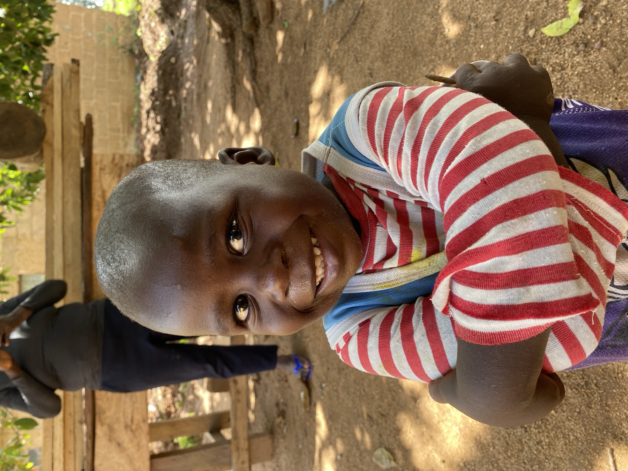 A medium shot depicts a smiling young boy in a red and white striped shirt, positioned with his body angled to the right. The boy is holding a stick in his hands. In the background to the left, a person in a dark shirt and dark pants is partially visible near a fence constructed from wooden planks. The environment suggests an outdoor setting, possibly a yard or open space, with dirt ground and scattered debris. Based on the location provided, the scene is in Riyom, Nigeria.