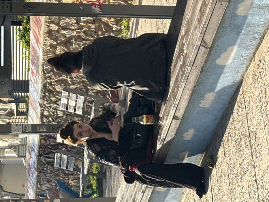 The image captures an outdoor scene in Tel Aviv, Israel, featuring two individuals seated on a long wooden bench. The bench's concrete base is painted light blue with white cloud motifs.
On the left, a female subject with reddish-brown hair styled in an updo is observed. She wears a dark, possibly lace or sheer, long-sleeved top over a dark garment and dark pants. She faces the viewer with a slightly open mouth, her right hand extended in a gestural motion towards the second subject, while her left hand holds a small, clear, cylindrical object. A dark fabric item, possibly a coat or bag, rests on the bench next to her.
On the right, a second individual is seen from behind, wearing a dark hooded jacket, with dark hair pulled into a top knot. This subject is facing and engaged in interaction with the first subject.
Between the two individuals on the bench, a small, clear plastic bottle with a green cap, containing a yellow liquid, is present. A pink smartphone is partially visible on the bench near the second subject.
The background features a stone wall with painted or graffiti-like markings in red, white, and blue hues. Behind this wall, green foliage, additional metal slatted benches, and parts of buildings with horizontal features are visible. To the far left, an older male subject with a receding hairline is seated on a light blue metal bench, facing away from the primary subjects. The ground consists of light-colored, textured paving stones. The scene is illuminated by strong, directional sunlight, casting distinct shadows. A dark metal post, partially covered with stickers and with some Hebrew text visible, stands in the foreground to the right.