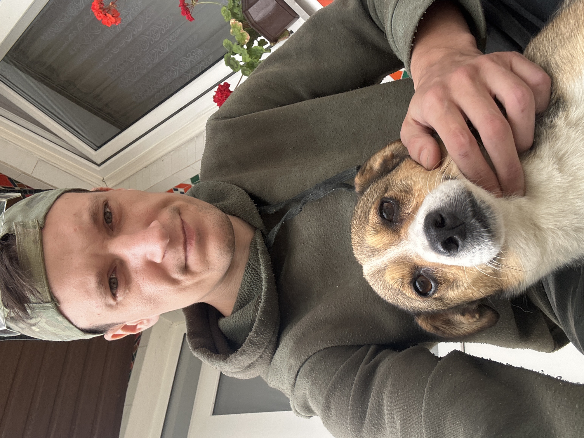 A man wearing a camouflage baseball cap and a dark green hooded sweatshirt is holding a small dog. The man's face is visible, with a slight smile. The dog, a mixed breed with brown and white fur, is looking directly at the camera. The man's right hand is gently resting on the dog's head. They are outdoors, with a white-trimmed window and red flowers in the background, suggesting a residential setting in Makiv, Ukraine. The image appears to be a selfie.