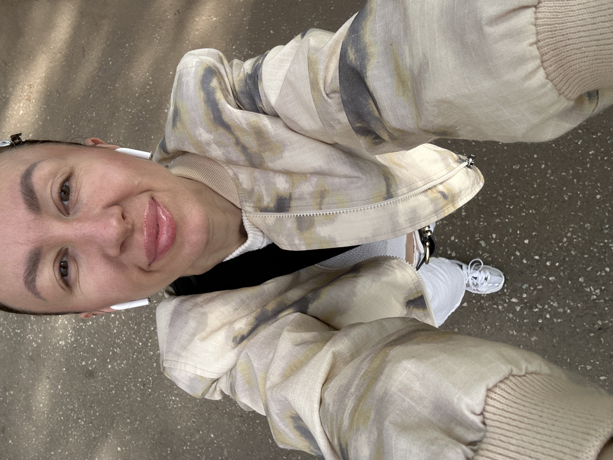 A woman is taking a selfie outdoors. She is wearing a light-colored jacket with a tie-dye pattern, a white shirt, and white sneakers. She also has white wireless earbuds in her ears. The ground appears to be paved. It is not possible to identify the location as Votkinsk, Russia, from the image alone.