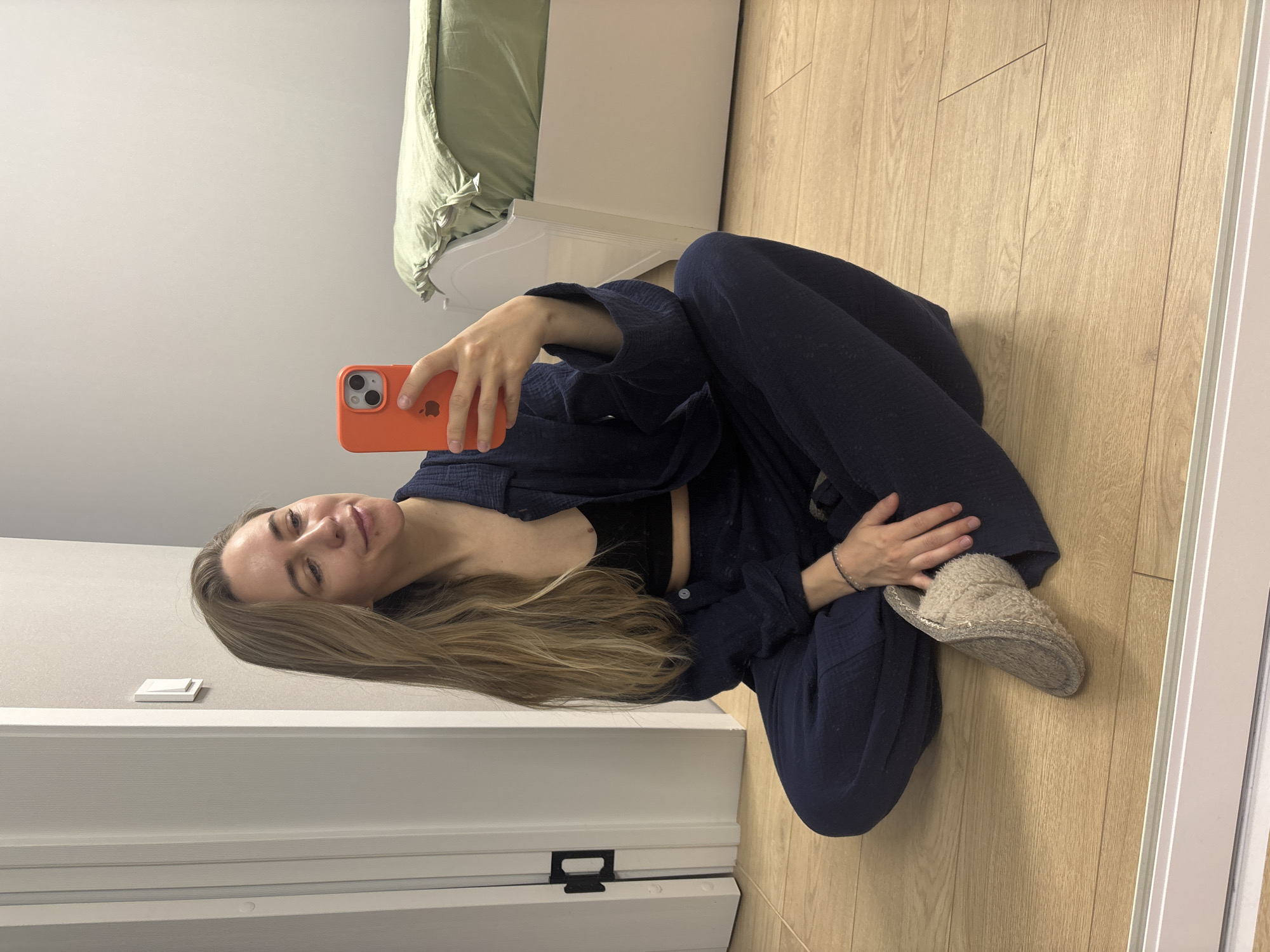 A young woman is captured in a selfie, sitting comfortably on a light wooden floor in an indoor setting in Chaikovsky, Russia. She is positioned with her legs bent, leaning slightly back, and a calm, soft smile on her face. Her long, light brown hair falls over her shoulders. She wears a relaxed, dark navy blue two-piece loungewear set, possibly made of textured cotton or gauze fabric. Beneath the unbuttoned top, a black bralette is visible. On her feet, she wears light beige, fluffy house slippers.
In her right hand, she holds a bright coral-orange smartphone, pointing it towards herself to take the photograph. The phone's back clearly displays a black Apple logo.
The background of the room is simple and light-toned. The floor is made of light-colored wooden laminate. A white wall extends behind her, featuring a white light switch. To her left, a white bed frame is partially visible, topped with a rumpled light green duvet or sheet, suggesting a bedroom environment. The lighting is soft and even, indicating an interior space, and the overall atmosphere is one of casual comfort and relaxation.