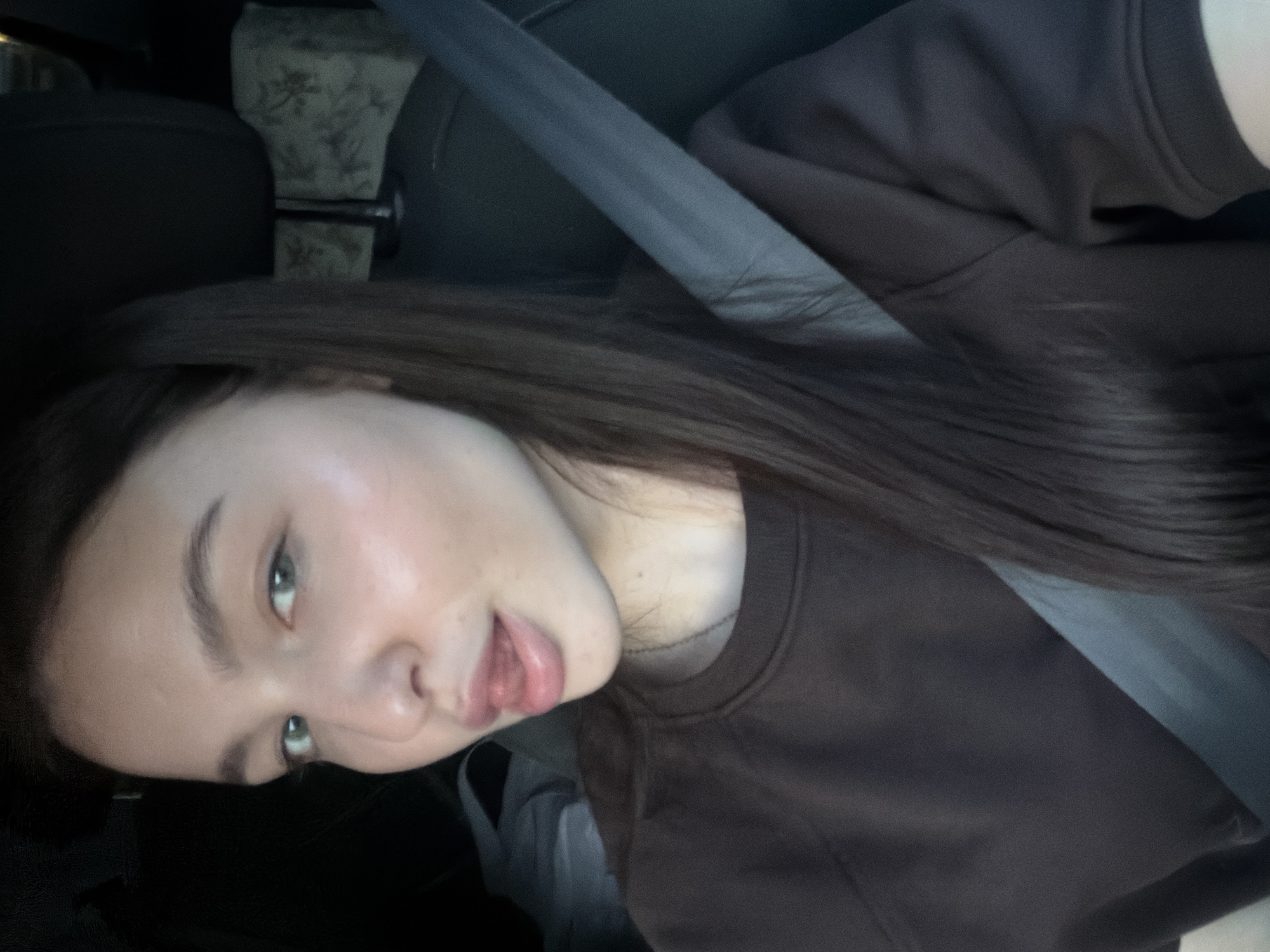 A young adult female is depicted in a close-up, chest-up shot from within a vehicle. She has fair skin, long dark brown hair falling over her left shoulder, and light green eyes. Her expression involves a direct gaze at the camera with a slightly open mouth, the tip of her tongue protruding. She wears a dark brown long-sleeved crew-neck top, with a thin metallic necklace visible. A gray seatbelt is fastened across her chest and right shoulder. Behind her, a dark car seat headrest is visible, and to her right (behind the headrest), a light-colored object with a faint floral pattern is partially seen. The lighting is soft and ambient. The scene is set inside the passenger compartment of an automobile.