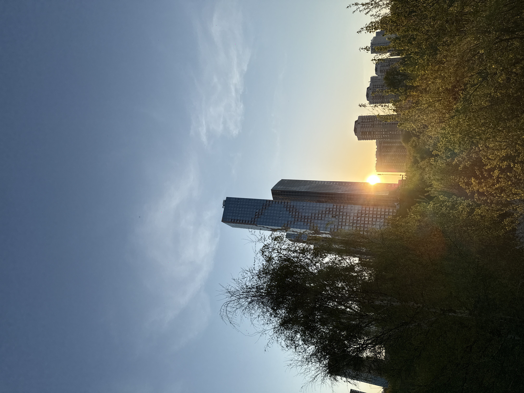 The image shows a view of Moscow, Russia at sunset. Two tall modern skyscrapers dominate the left side of the frame, while a row of residential buildings can be seen in the distance on the right. The sun is setting between the buildings, creating a bright glare. Trees frame the right side of the image. The sky is a light blue with some wispy clouds. The photo is rotated.