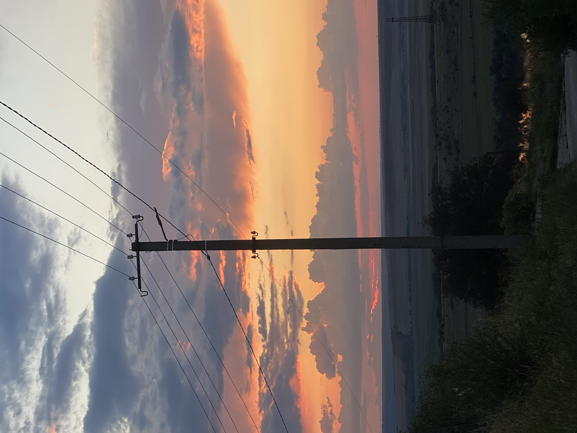 This is a photograph taken in Cairaclia, Moldova, at sunset. The image is dominated by a dramatic sky filled with clouds painted in hues of orange, pink, and purple, transitioning to a pale yellow and gray near the horizon. A single, tall, concrete utility pole stands prominently in the foreground, slightly off-center to the right, with multiple electrical wires stretching across the frame from it.
Below the sky, a vast, open landscape unfolds. It appears to be a rural area with rolling hills and fields, some of which are darker green and appear cultivated, while others are a lighter, more muted tone. In the distance, there are hints of more hills and possibly some structures, including a tall, lattice-like transmission tower on the far right. The foreground is darker, with the silhouettes of trees and tall grass. A small body of water, possibly a river or pond, is visible in the lower right quadrant, reflecting some of the sunset light. The overall impression is one of a peaceful, rural evening scene.