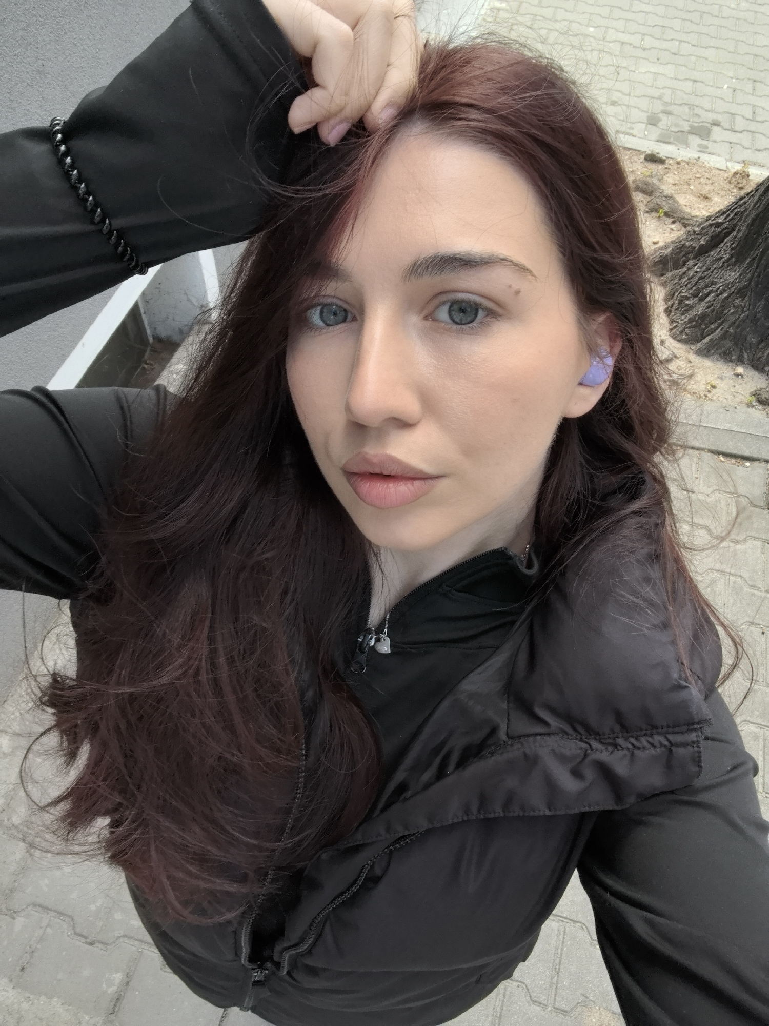 A woman is taking a selfie outdoors. She has long auburn hair, blue eyes, and is wearing a black puffy jacket, a black shirt, and a beaded bracelet. She has a purple earbud in her left ear and is touching her hair with her left hand. The background shows a grey paved surface, possibly a sidewalk, with a tree trunk and foliage in the upper right corner. Based on similar images, this is located in Stara Zagora, Bulgaria.