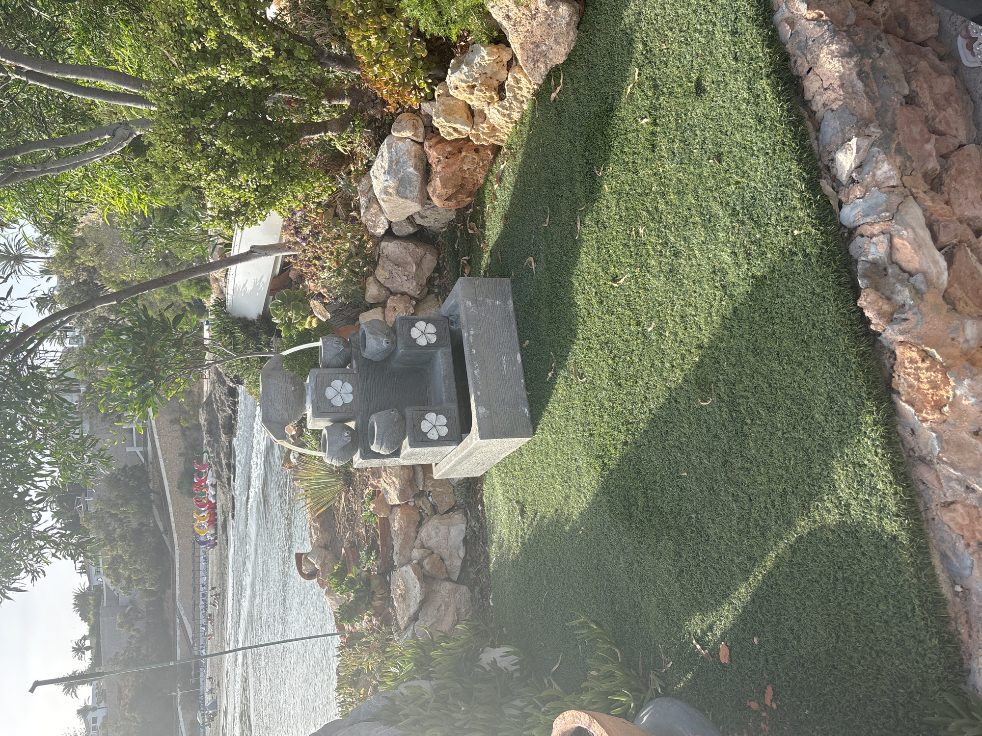 A stone fountain with multiple levels and flower carvings sits on a patch of artificial grass. The fountain has water trickling down its tiers. Behind the fountain, rocks of various sizes are arranged, interspersed with plants and small potted succulents. Beyond the rocks, a body of water is visible, with a beach lined with sun loungers and colorful paddle boats. In the background, a hillside is covered with trees and buildings. The scene appears to be outdoors, likely in a coastal area.