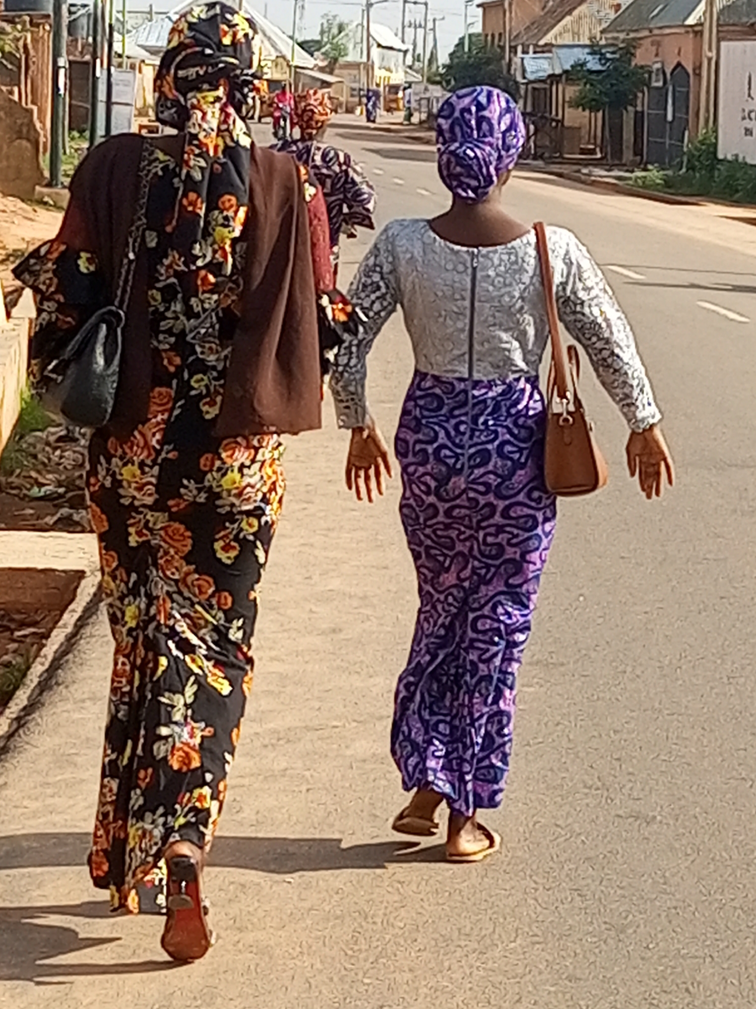Two women are walking away from the camera on a paved road in Gombe, Nigeria. The woman in the foreground is wearing a dark brown jacket over a black dress with a vibrant floral pattern in orange, yellow, and white. She has a black handbag slung over her left shoulder and is wearing red high-heeled shoes. Her head is covered with a patterned headscarf that matches her dress. To her right, a second woman is wearing a light-colored, possibly silver, lace top with a zipper down the back. Her long skirt is a deep purple with an intricate dark purple pattern. She is carrying a tan handbag on her right shoulder and is wearing light-colored flat sandals. Her head is covered with a purple and white patterned headscarf. In the background, there are buildings with reddish-brown roofs and shops lining the street. The road has white lane markings, and there is a pedestrian walkway to the left of the women. The scene appears to be a sunny day.