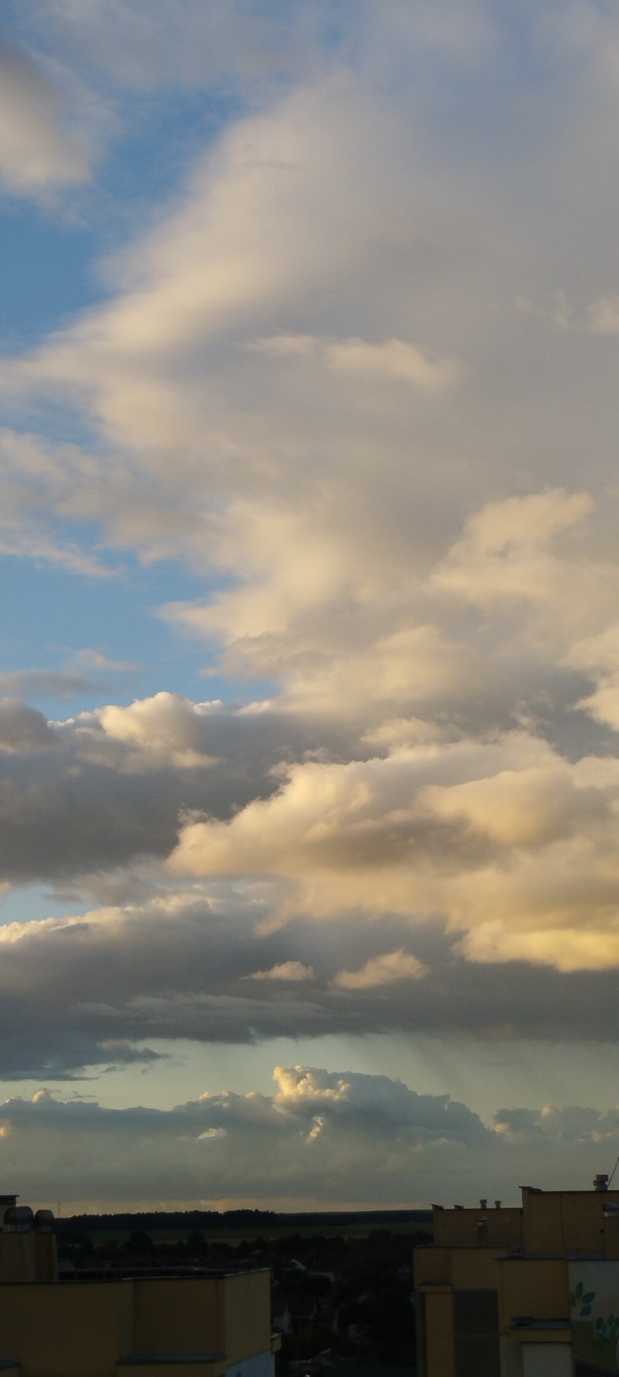 The image depicts a cloudy sky over a cityscape, likely Skidzieĺ, Belarus. The clouds are a mix of white and gray, with hints of yellow and orange, suggesting either sunset or sunrise. The lower part of the image shows the rooftops of several buildings, some of which appear to be apartment blocks. A dark line of trees is visible in the distance, separating the city from the horizon. The overall lighting suggests it is late afternoon or early evening. There are no people or animals visible in the frame. The composition is dominated by the sky, with the cityscape serving as a backdrop.