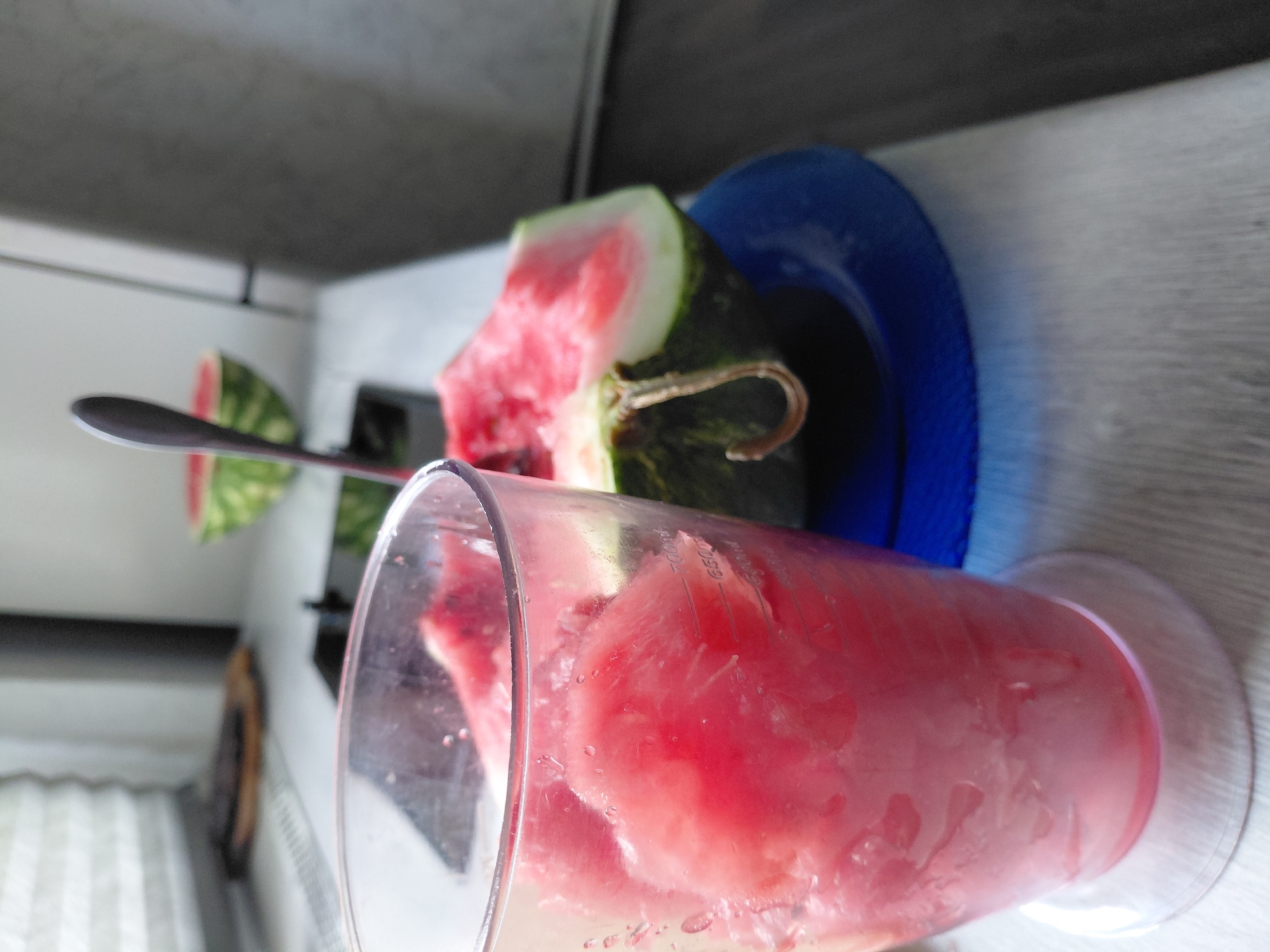 A clear plastic cup filled with pink watermelon chunks sits on a white countertop. A blue saucer rests beneath the cup. In the background, a slice of watermelon with a green rind and red flesh is visible on the saucer, with a silver spoon inserted into it. The setting appears to be a kitchen, likely in Novosibirsk, Russia, based on the muted lighting and common kitchen appliances visible in the background. The scene suggests preparation for making a watermelon smoothie or drink.