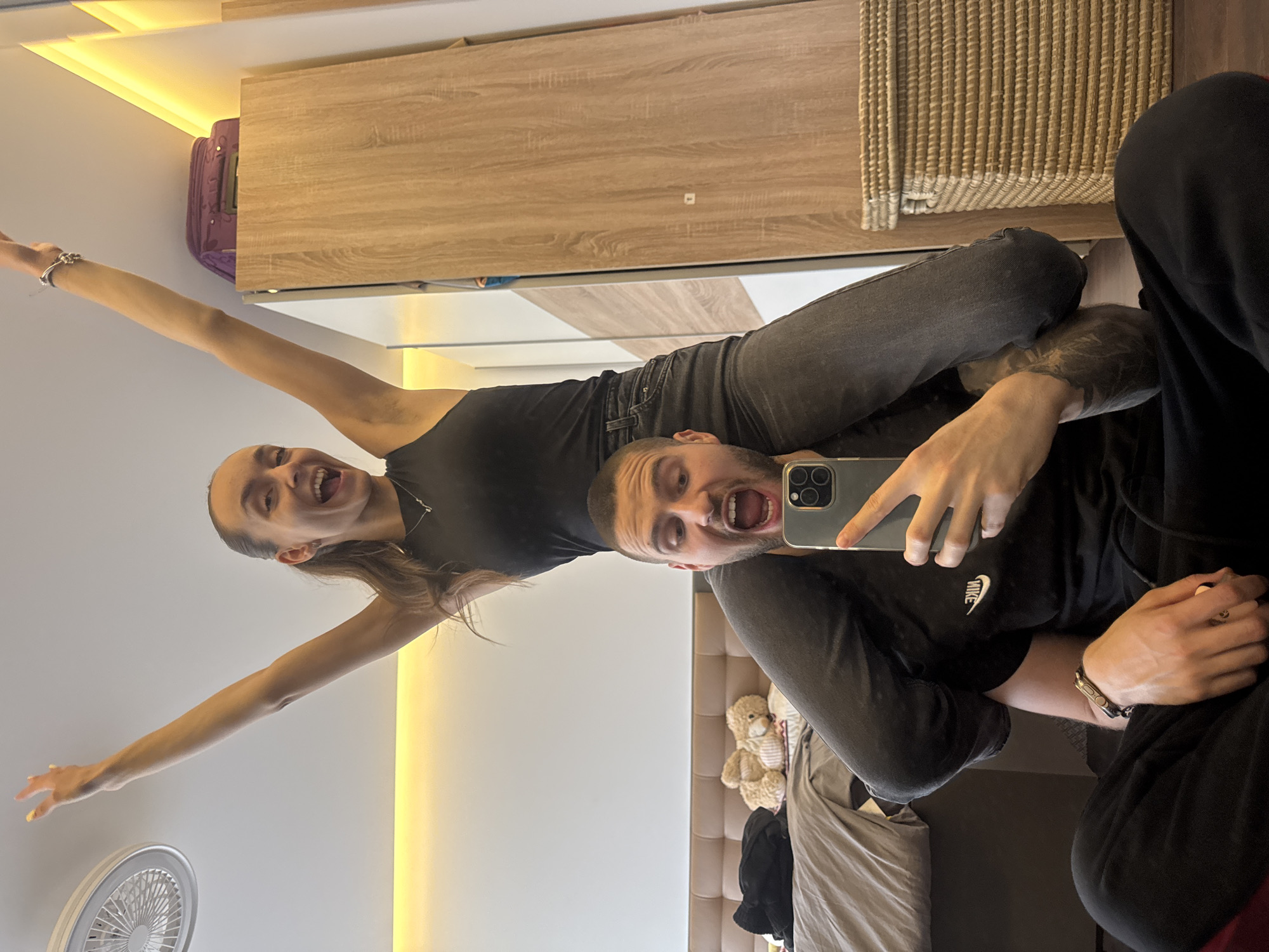 A man and a woman are posing for a photo in what appears to be an apartment interior. The man is in the foreground, holding a smartphone to take a selfie. The woman is behind him, on his shoulders, with her arms raised. She is smiling broadly. The man is looking at the camera with his mouth open.
The background shows a room with light-colored furniture, a fan, and a bed. The location cannot be determined from the image.
