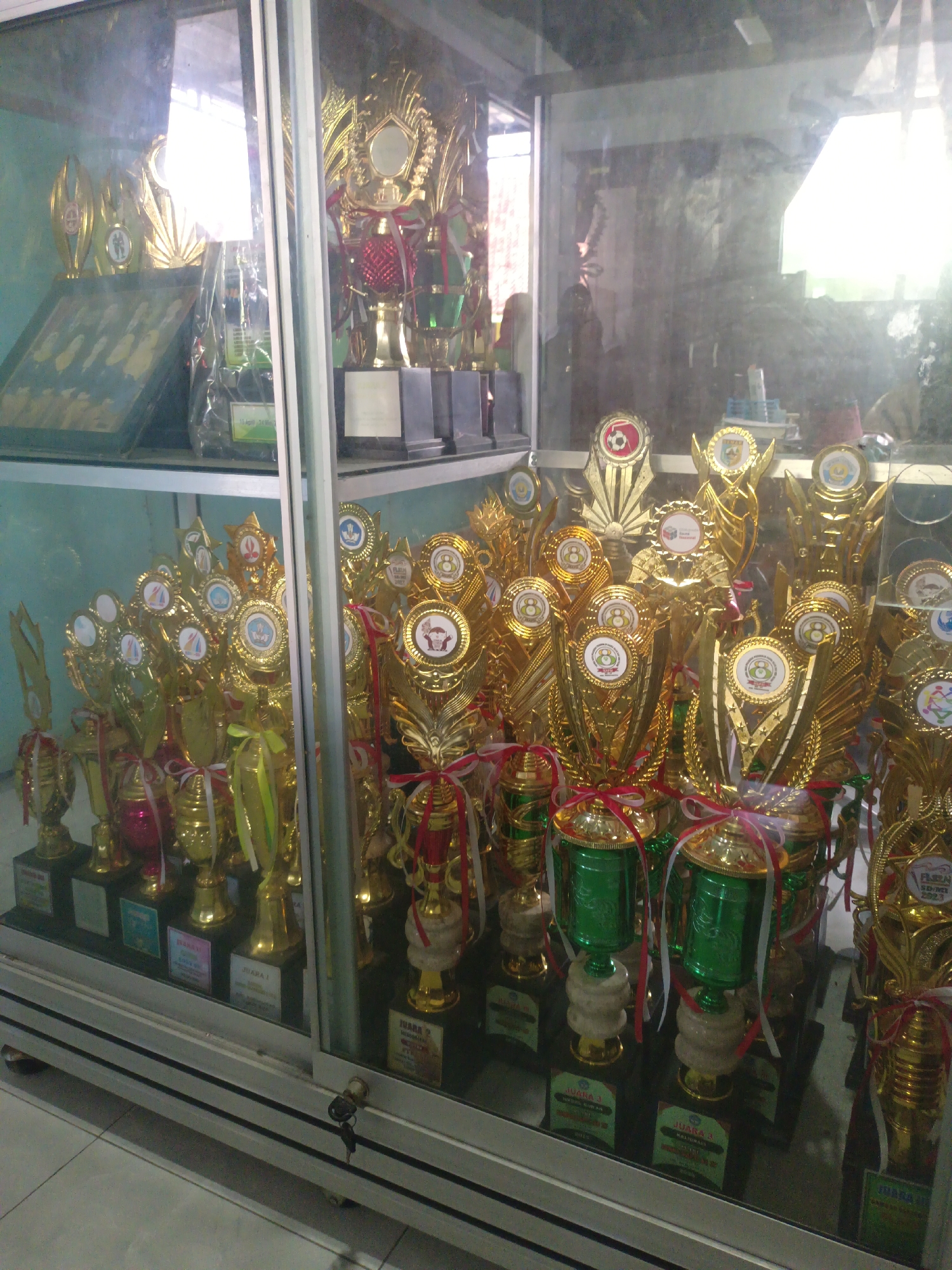 The image shows a display case filled with numerous gold and green trophies, likely for sports or academic achievements. The trophies vary in size and design, with many featuring circular emblems at the top. Some trophies are adorned with red and white ribbons. The display case appears to be made of glass and metal, with a key in the lock at the bottom. The background is blurred, but suggests an indoor setting with windows and reflections of the outdoors. The setting is likely a school or community center in Sarimulyo, Indonesia, given the context provided.