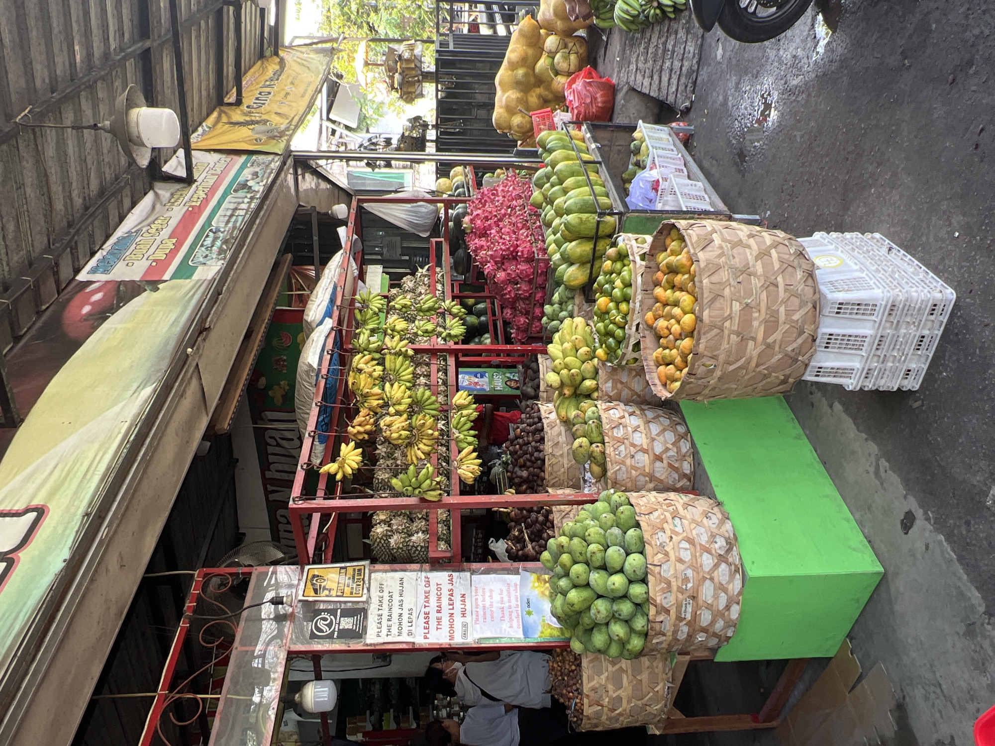 This image captures a vibrant fruit stand, likely in North Kuta, Indonesia. The stall is brimming with a colorful assortment of fruits including bananas, mangoes, dragon fruit, oranges, and pears. The fruits are arranged in woven baskets and on shelves within a red metal frame. Signs instruct customers to remove raincoats before entering the shop, indicating frequent rain in the area. Beneath the stall's signage, two individuals are partially visible, possibly customers or vendors. The surrounding environment includes a paved street and the edge of a motor vehicle. The overall scene depicts a bustling local market offering fresh produce.