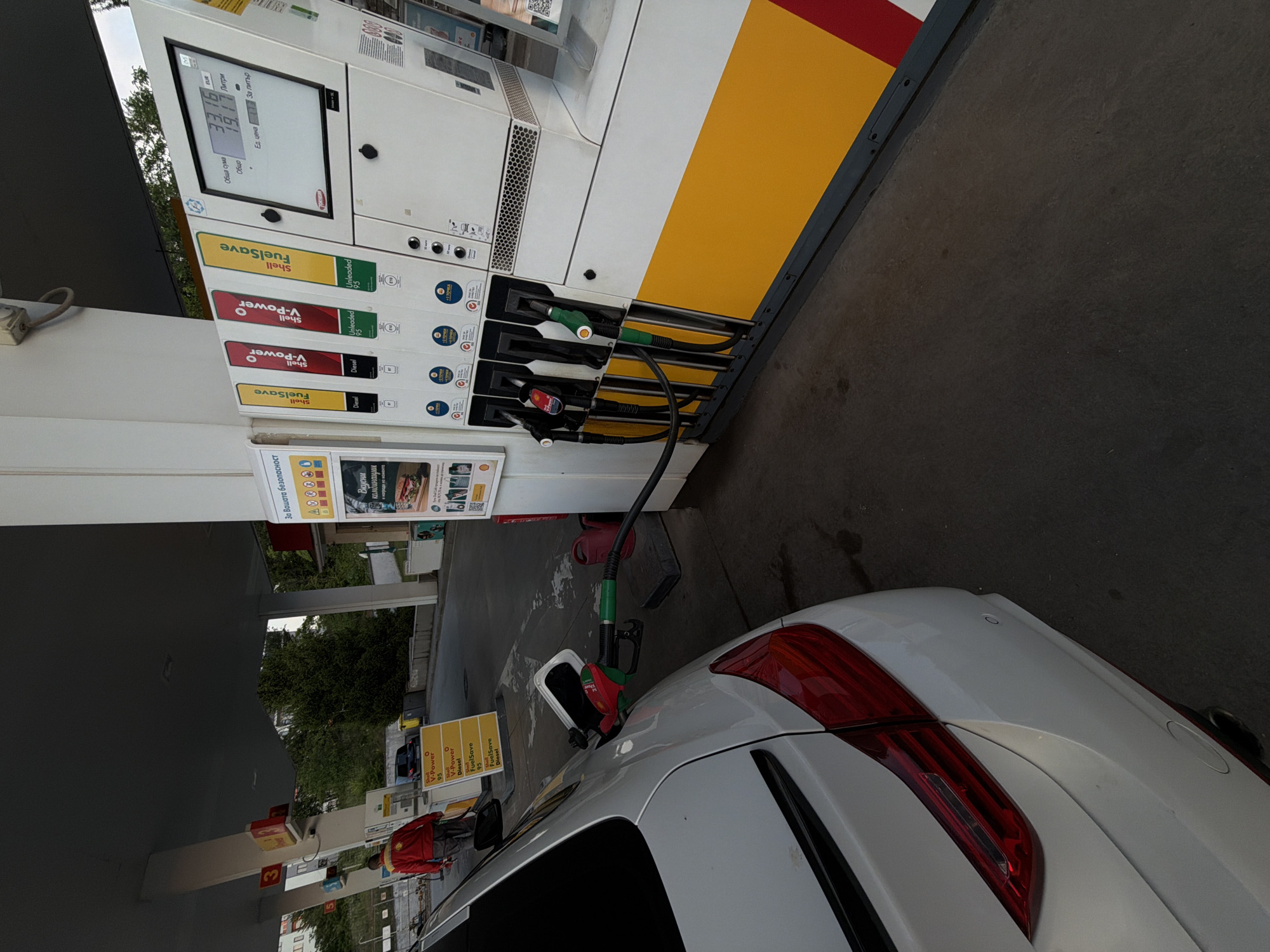 A white car is seen from the rear, being refueled at a Shell gas station. A green and red fuel nozzle is inserted into the car's fuel tank, while the hose extends back to a multi-product fuel dispenser. The dispenser features the Shell logo and displays various fuel types, including "Shell FuelSave Unleaded 95," "Shell V-Power Unleaded 95," "Shell V-Power Diesel," and "Shell FuelSave Diesel."
The digital screen on the fuel pump shows the transaction details. The "Обща сума" (Total amount) is 33.16 EUR, and "Общо" (Total) "Литри" (Liters) dispensed is 19.39. Below these figures, "Ед. цена" (Unit price) and "За литър" (Per liter) are visible. Text on a sign below the pump reads "За Вашата безопасност" (For your safety), indicating the language is Bulgarian.
The setting appears to be daytime, with a gas station canopy providing overhead cover. In the background, trees and other structures are visible, along with another car parked further down. A person in a red jacket is faintly visible near other fuel pumps. The ground beneath the pump and car is concrete.