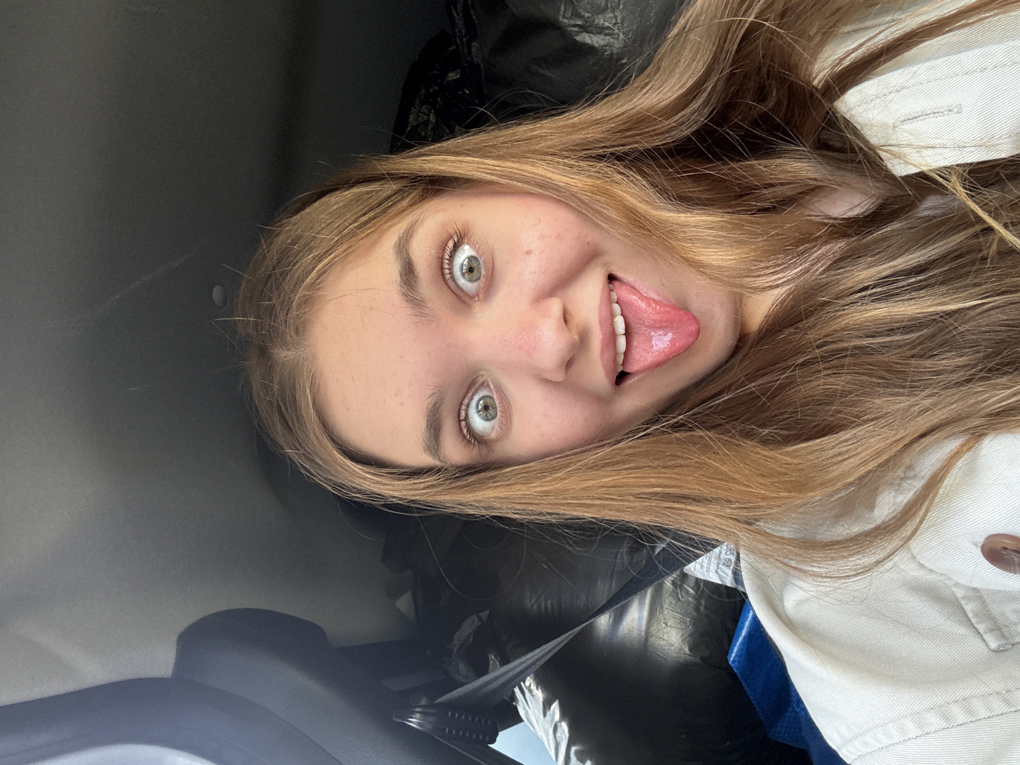 A young woman with fair skin and long, light brown hair sticks out her tongue at the camera. She has green eyes. She is wearing a white jacket. The background is partially obscured, but it appears she may be sitting in a car. There is a black plastic bag visible behind her. There is no evidence that the image was taken in Perm, Russia.