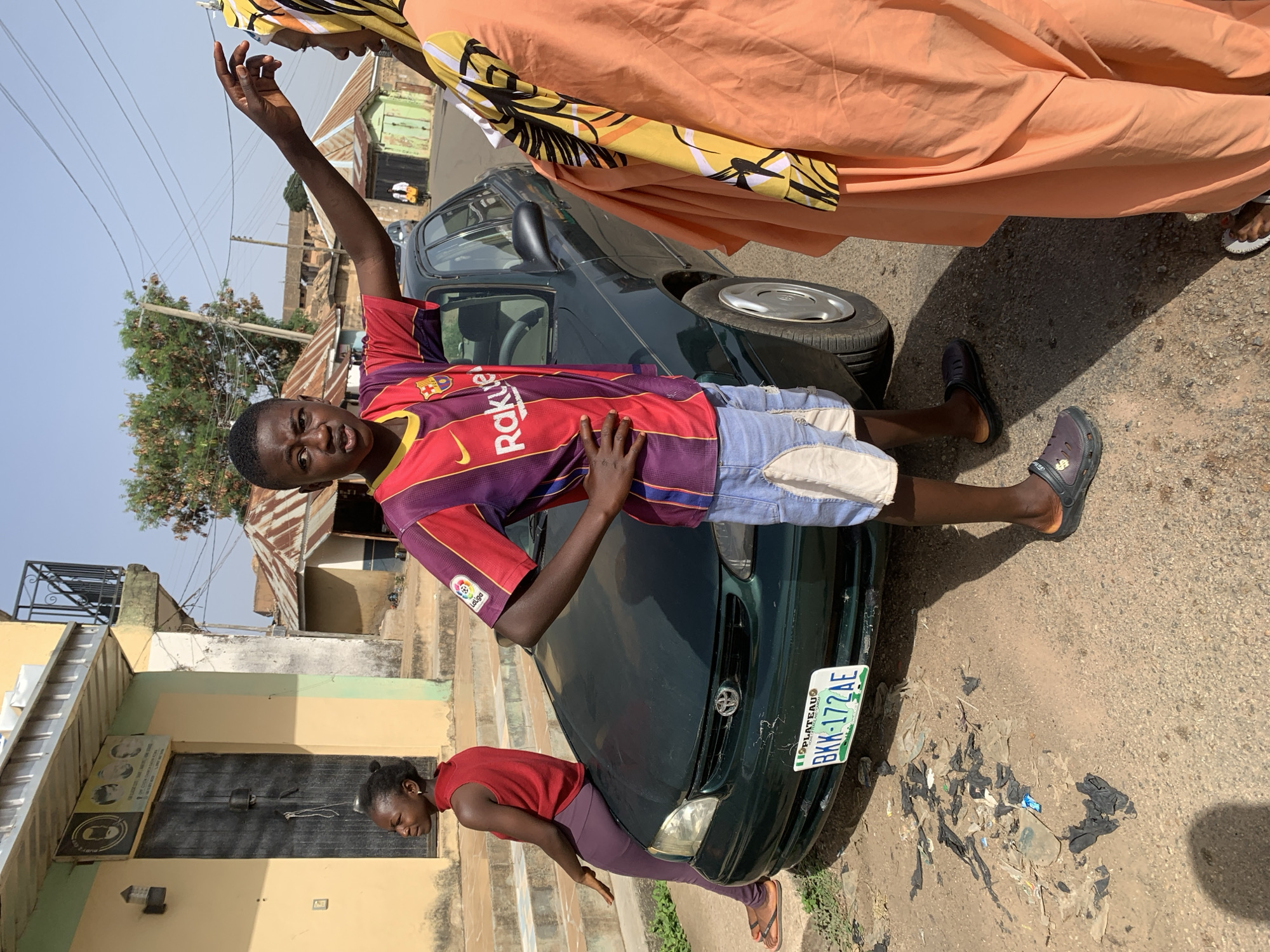 A teenage boy in a red and purple Barcelona soccer jersey and light blue shorts is posing on a dark green Toyota car with a Nigeria Plateau license plate "BKK-172 AE". He has one arm raised and the other on his hip. Another woman is crouched down to the left of the car wearing a red tank top and purple pants. Another person in peach clothing stands next to the car. The scene is an urban neighborhood with buildings and trash-strewn streets.