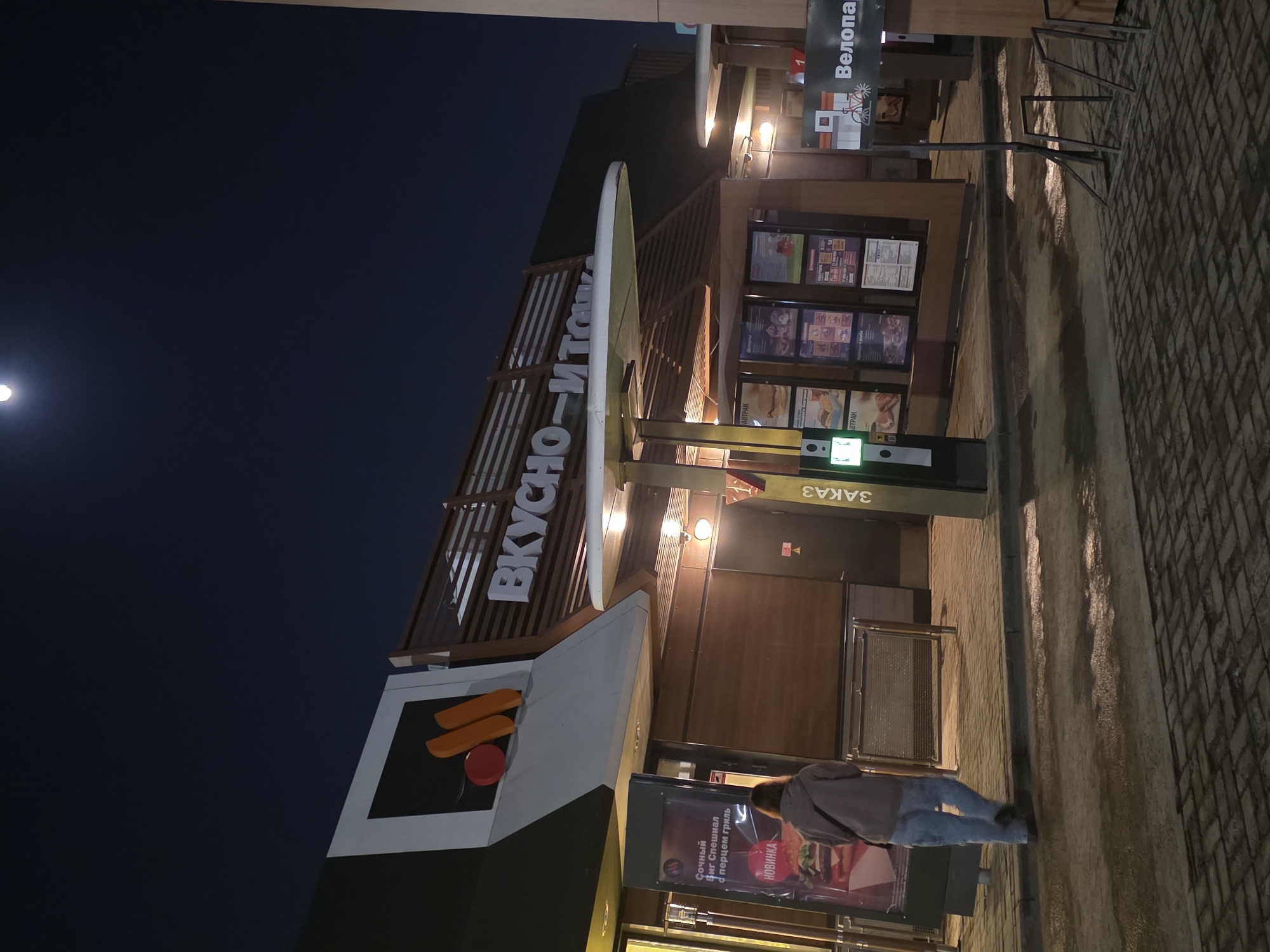Here is a detailed description of the image:
**Scene:** The image shows a McDonald's restaurant in Izhevsk, Russia. The restaurant's new name, "Вкусно и точка" (Vkusno i tochka), is visible on the building. The sky is dark, indicating it is either evening or night.
**People:** A person wearing a hoodie and jeans stands in front of the restaurant, seemingly looking at the advertisement displayed in the window.
**Objects:**
* The restaurant's signage, including the "Вкусно и точка" logo.
* A promotional advertisement is displayed in the window.
* A "ЗАКАЗ" (Order) sign.
* A bicycle rack.
* Menu boards displaying food items.
**Environment:**
* The restaurant is located on a paved street, potentially in a commercial or urban area.
* The lighting suggests it is either early morning or late evening.
* The overall atmosphere appears calm.