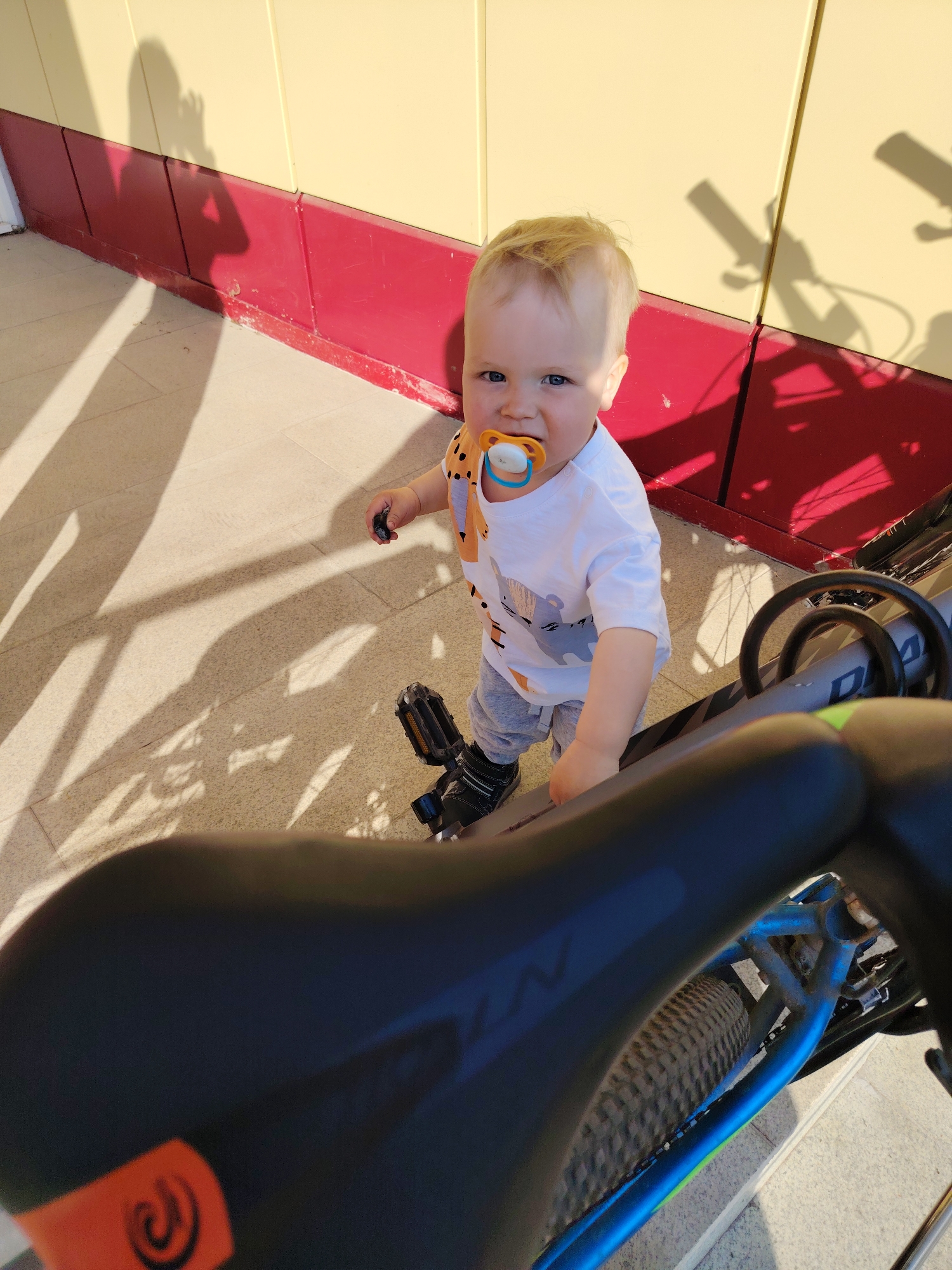 A fair-skinned baby with blonde hair and blue eyes stands outdoors, holding a small black object in their left hand. The baby is wearing a white t-shirt with gray pants, and a pacifier is in their mouth. The child is next to a bicycle, standing on one of the pedals, with the seat and frame of the bike prominently in the foreground. The bike has a blue and gray frame with black tires. The location appears to be a paved area outside a building with a yellow and red wall. Shadows are cast on the ground, likely from the sun. Based on the user's information the location is Chaikovsky, Russia.