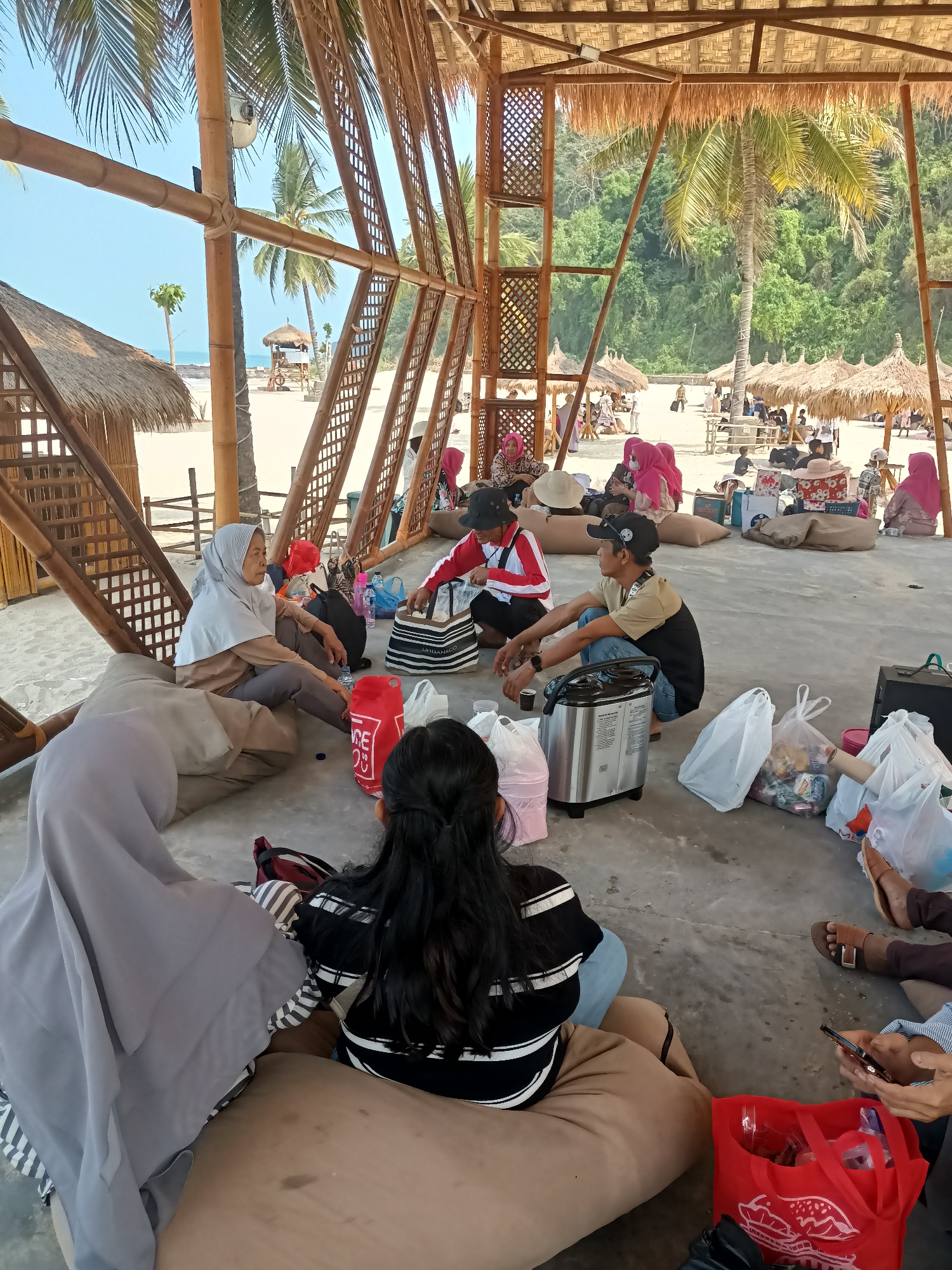The image depicts a group of people gathered in an open-air bamboo structure near a beach, likely in Bulok, Indonesia. The structure, with its woven bamboo walls and thatched roof, provides shade. In the foreground, several individuals are seated on large floor cushions or beanbags. Some are wearing headscarves, indicating a cultural context.
Several objects are scattered around the group, including a silver bread maker, plastic bags filled with items, and a large red tote bag with a graphic print. One person in the foreground is holding a smartphone.
The background reveals a sandy beach with palm trees and a clear blue sky. Further back, there are more bamboo structures with thatched roofs and a scattering of people. The overall scene suggests a relaxed atmosphere, possibly a gathering or a stop during a trip to the beach. The presence of the bread maker implies that food preparation might be part of their activity. The environment is tropical, with lush green foliage visible on a hill behind the beach.