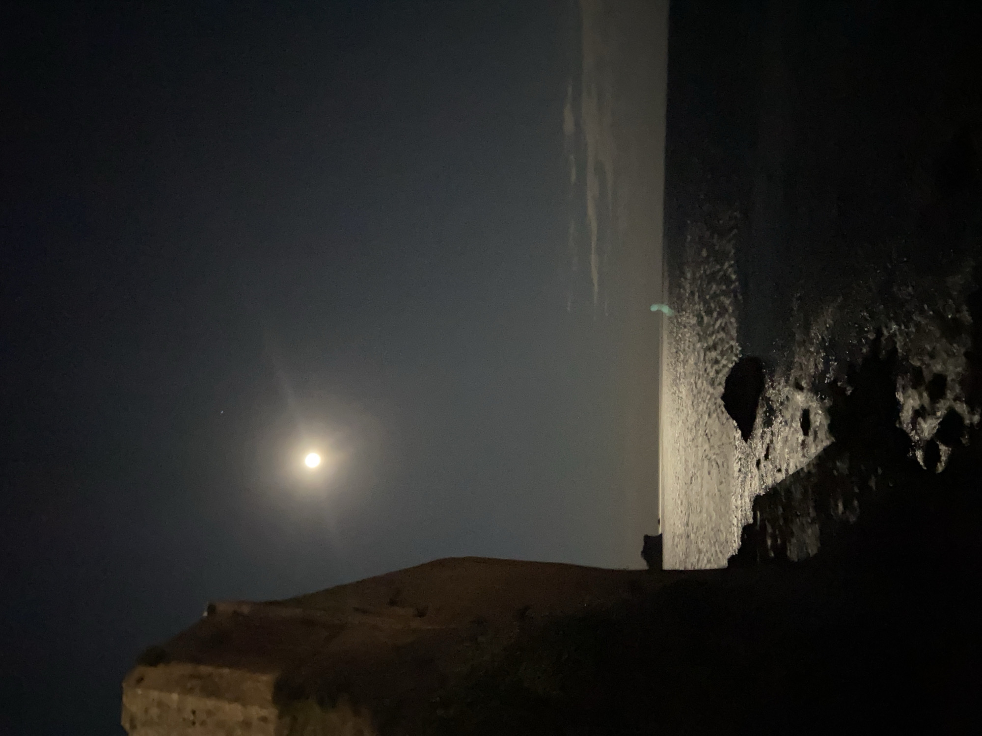 The image captures a serene nighttime coastal landscape, likely in Budva, Montenegro. The scene is dominated by a bright, nearly full moon positioned in the upper left quadrant, casting its luminous glow across the dark sky and shimmering sea. A small, bright celestial body, possibly a star or planet, is visible slightly above and to the right of the moon.
The sky is a deep indigo-black, with faint, wispy clouds visible in the upper right. A clear horizontal line, the horizon, separates the dark sea from the distant sky or a hazy coastline in the middle-right of the frame. The sea in the lower right portion of the image reflects the moonlight, creating a textured, silvery appearance on its surface, indicating gentle waves or ripples. A faint, almost neon green light source is discernible near the horizon line over the water, possibly a distant buoy or boat light.
In the foreground, occupying the lower left and central portions of the image, is a dark, rugged landmass, appearing as a cliff or hillside. Its contours are dimly lit, suggesting rough terrain. At the very bottom left, part of a substantial stone structure, possibly an old wall or fortification, is visible, adding a historical or architectural element to the natural night scene.
The time of day is clearly night, illuminated primarily by the moon. The overall atmosphere is peaceful and dramatic, emphasizing the vastness of the night sky and the sea under moonlight. There is no visible text or human activity depicted.