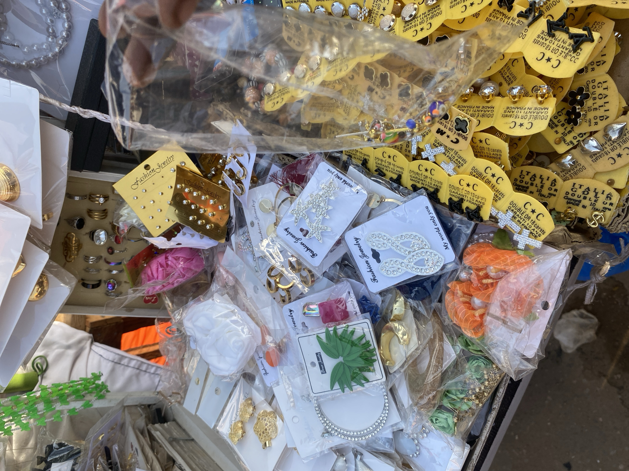 The image presents a close-up, high-angle view of a market stall displaying a variety of fashion jewelry and accessories in Sabon Tasha, Nigeria. The majority of the merchandise is individually packaged in clear plastic sleeves, often affixed to white or yellow cardboard backing cards, many labeled "Fashion Jewelry" or "Benze Jewelry."
The stall is densely packed with items, including various earrings (studs, hoops, dangling designs, some with cubic zirconia), necklaces (a string of silver-colored pearls is visible), bracelets (a large, circular silver-colored piece with embedded stones), and rings. Hair accessories are prominent, featuring orange sheer fabric scrunchies, white floral clips, green leaf-shaped clips, and silver-colored star-shaped and infinity-loop clips adorned with stones.
A large quantity of small pendants and charms are presented on distinct yellow, C-shaped tags. These tags bear text such as "DOUBLE OR 18 CARATS," "GARANTI 10 ANS," "MADE IN FRANCE," and "ITALIA GOLD." The attached charms include crosses, four-leaf clover shapes (some black, some with stones), heart shapes, and various small metallic emblems.
A dark-skinned hand is visible on the right side of the frame, holding a clear plastic bag filled with additional merchandise. In the background, partially obscured elements include a white garment, a bright orange object, and a green beaded garland. The items are arranged on what appears to be a flat display surface, with a dark ground surface visible at the bottom of the frame.