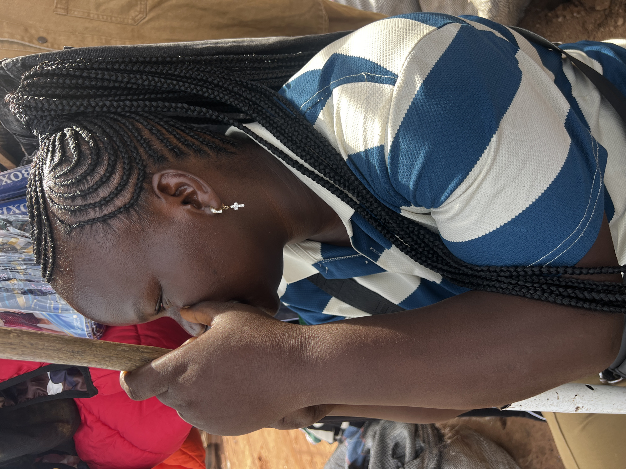 The image depicts a Black woman with braided hair, wearing a striped blue and white shirt and a small cross earring. She appears to be resting her head on her hand, which is holding a wooden stick. She is situated among various items, including a red puffy jacket and boxes. The location is in the city of Unknown, Unknown.