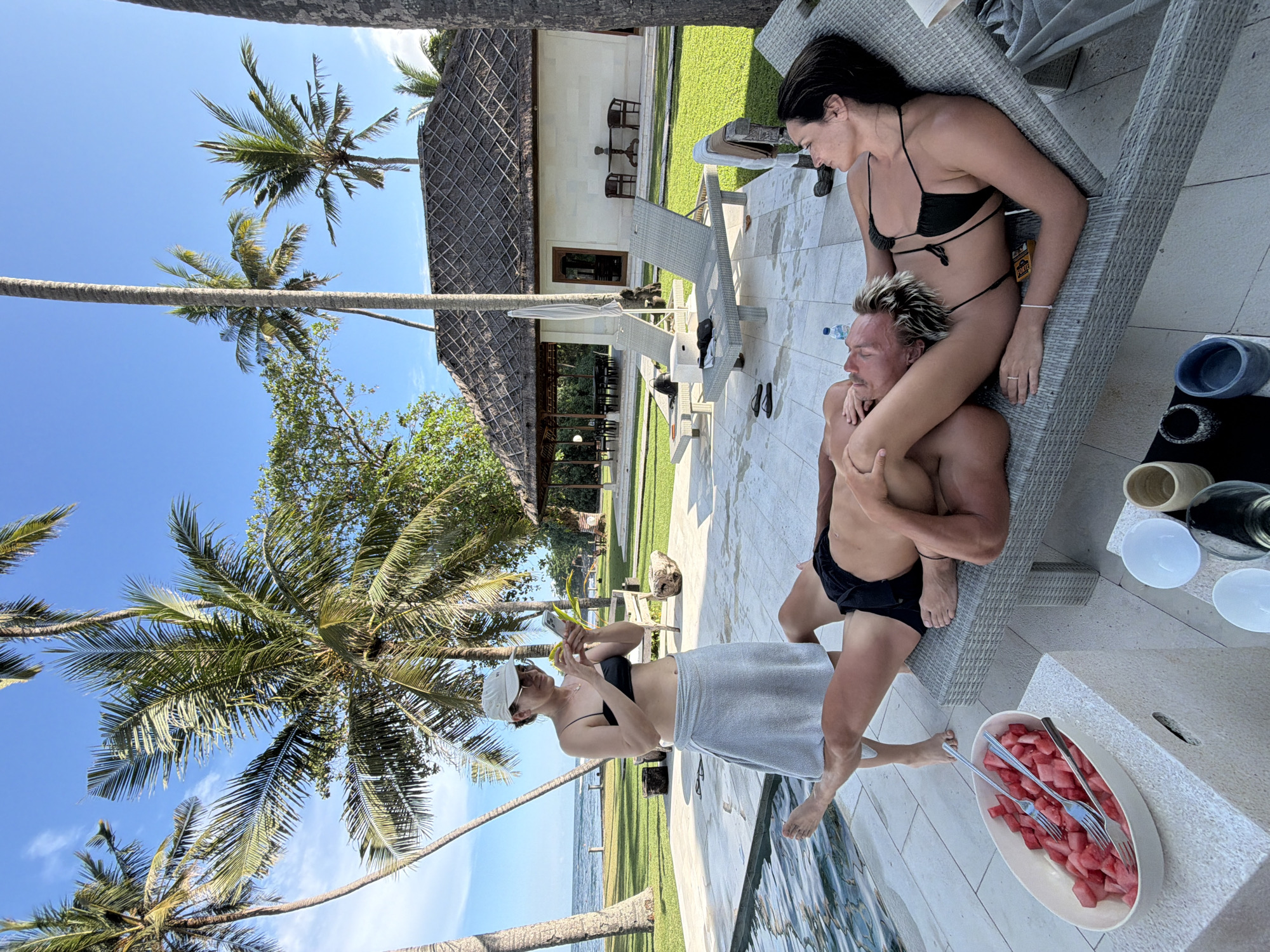 The image captures a scene at a resort in Karangasem, Indonesia. Three people are present. A woman in a black bikini sits on a wicker lounge chair, with a man in black swim trunks positioned beneath her, as if supporting her with his body. Another woman, wearing a black bikini top, a white baseball cap, and a grey towel wrapped around her waist, stands nearby and appears to be taking a photograph with her phone. A plate of watermelon with forks is positioned next to the lounge chair. The background includes palm trees, a view of the ocean, a building with a thatched roof, and manicured lawns.