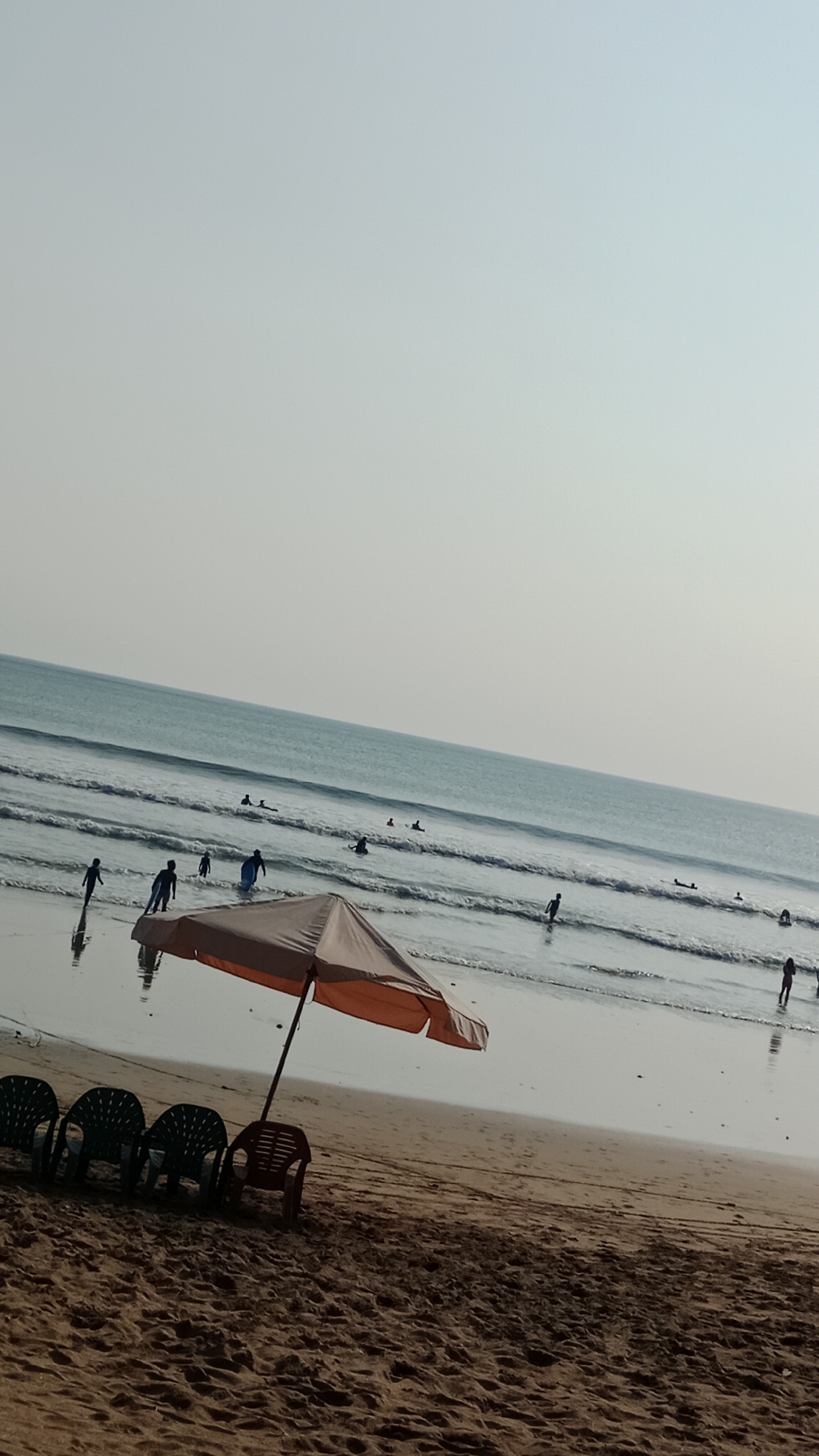 The image depicts a beach scene in Kuta, Indonesia. The foreground shows a sandy beach with several plastic chairs lined up and an open umbrella. In the midground, the ocean waves are breaking, and several people are visible in the water, some of whom appear to be surfing. The sky is a light, hazy blue, suggesting it might be late afternoon or early morning. The overall atmosphere is relaxed and recreational, typical of a beach resort.