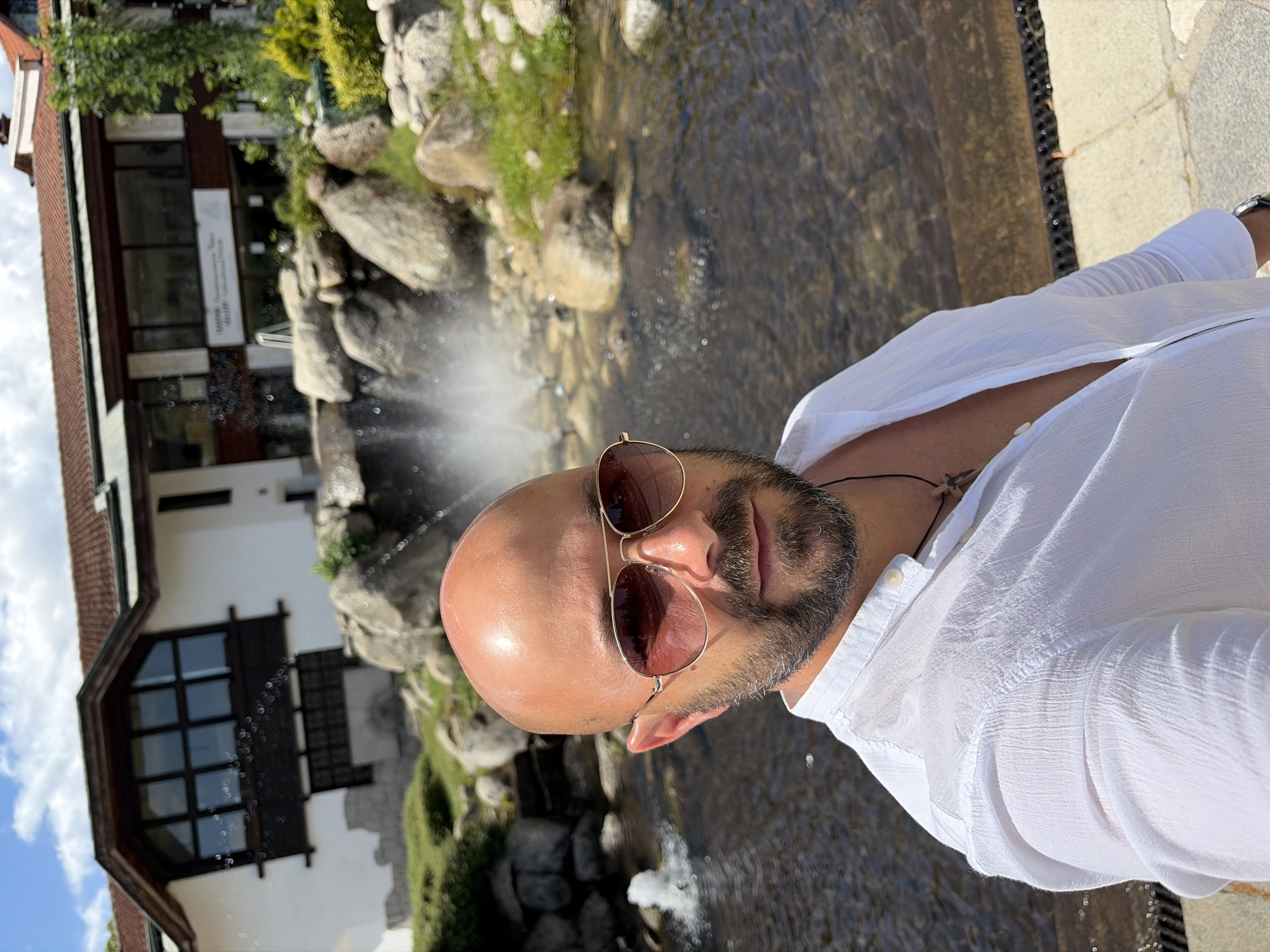 A man with a shaved head, sunglasses with rose-tinted lenses, and a beard is standing in front of a body of water with a waterfall in the background. He is wearing a loose-fitting white linen shirt. Behind him is a building with a red-tiled roof and large windows, characteristic of mountain architecture. The area appears to be a resort or hotel, possibly with a man-made water feature. Based on the context, this scene is likely located in Bansko, Bulgaria, a popular mountain resort town.