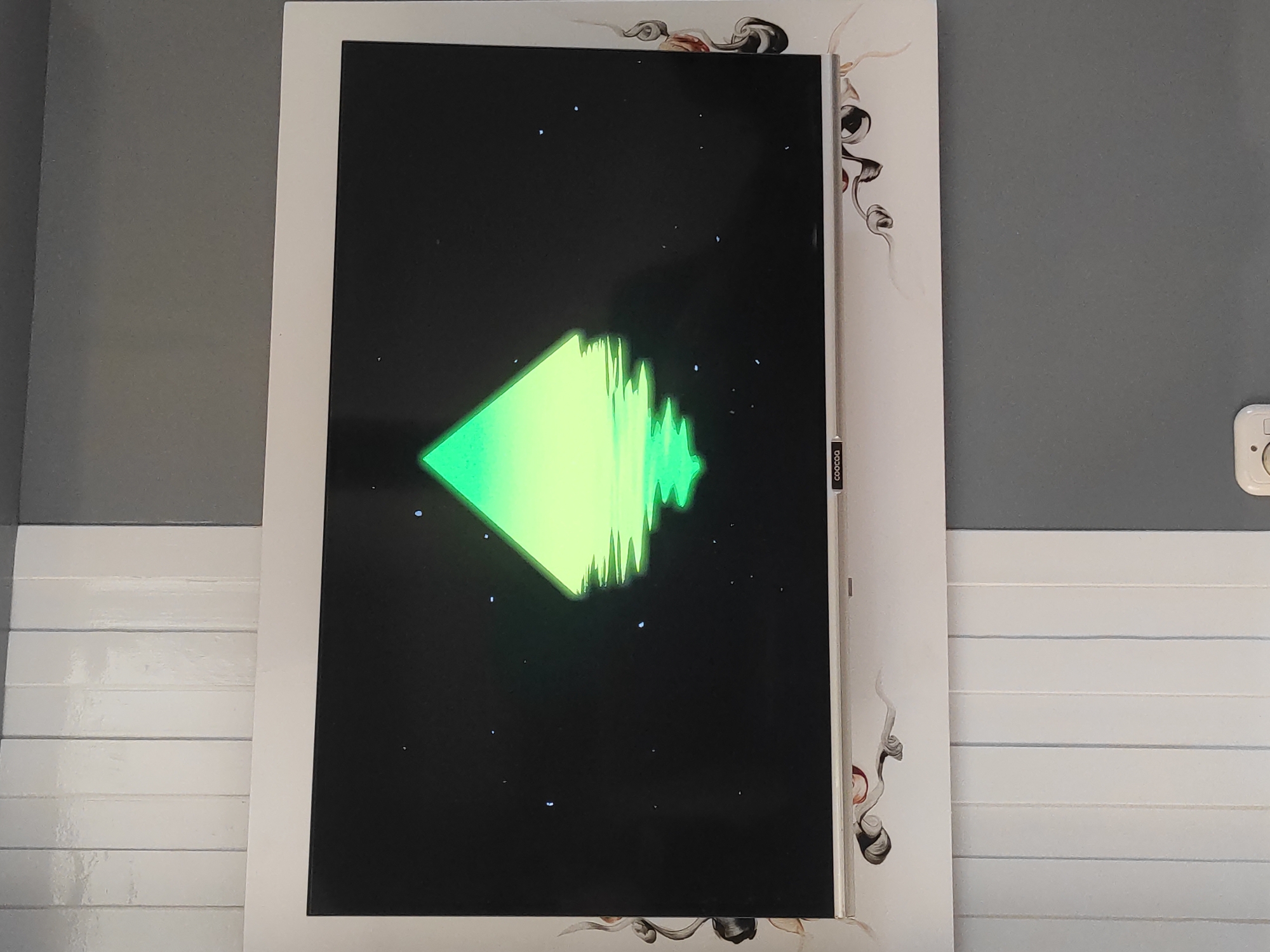 The image shows a flat-screen television mounted on a wall. The television displays a neon green pyramid shape with a reflection on a dark, starry background. The wall behind the television is painted in two colors: white vertical paneling on the left and grey on the right. Decorative swirling brown and white designs are applied to the wall around the television frame. The television has a silver bezel and a small logo, likely a brand name, is visible in the bottom left corner of the screen. The image is taken from a slightly elevated angle, looking down at the television. The scene appears to be indoors, likely a room in a residence or a public space. There is no indication of the city of Sumedang, Indonesia, in the image.
