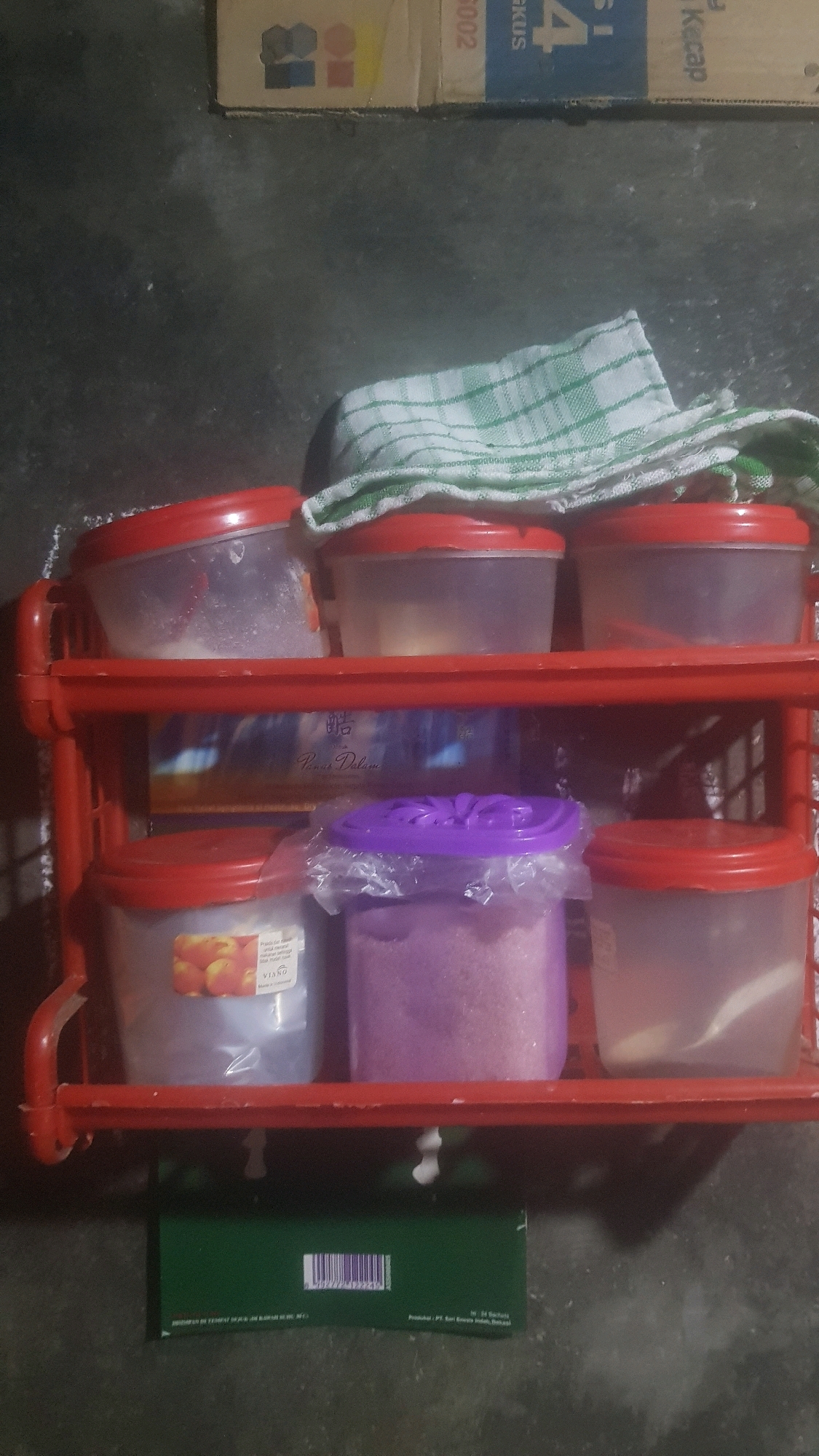 An overhead shot captures a red plastic two-tiered shelf unit holding six clear plastic containers with red lids and one container with a purple lid. The shelf unit is positioned on a dark, rough surface that appears to be concrete. Behind the shelf, on the upper left, is a portion of a cardboard box with colorful geometric designs and some text. A green and white checkered cloth is draped over the top of the shelf unit. The bottom shelf contains three containers: one with an image of peaches on its label, a second with a purple lid containing purple granular material, and a third holding a white granular substance. The top shelf holds three containers, each with a white granular substance. A green rectangular box with a barcode is placed on the dark surface below the shelf. The scene is indoors.
