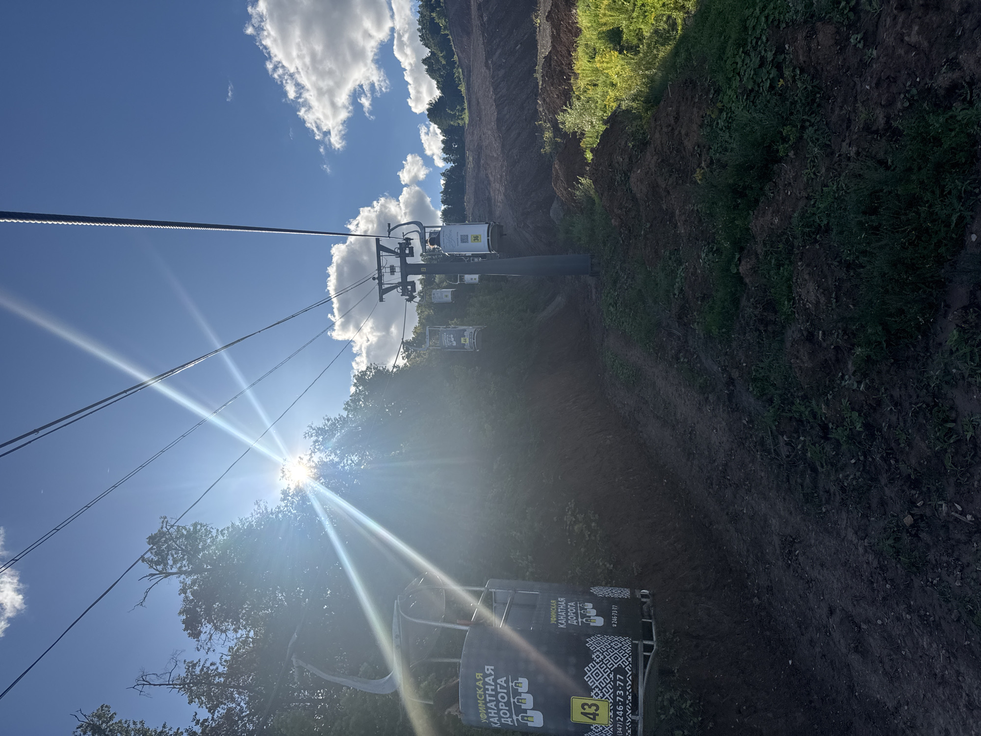 The image shows a cable car system in the city of Ufa, Russia. The cable car cabins are black and white with Cyrillic text that translates to "Ufa Ropeway". One cabin in the foreground is labeled with the number 43 and a phone number, (347) 246-73-77. The cable cars are suspended over a wooded hillside with a large area of disturbed earth and vegetation. The sun is shining brightly, creating lens flare across the image. The sky is blue with scattered white clouds. The scene appears to be a tourist attraction or recreational area.