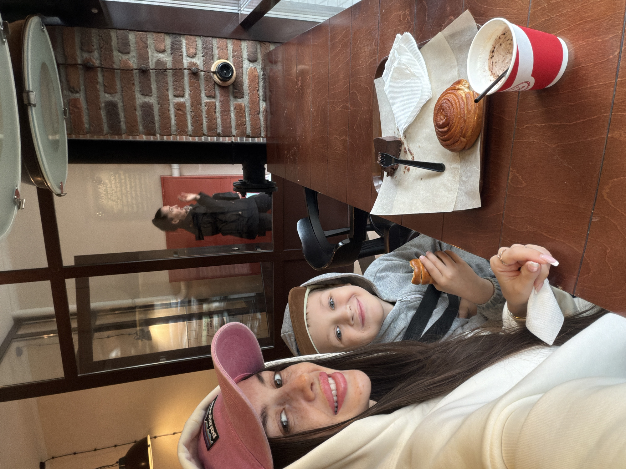 A woman in a pink baseball cap and a child are taking a selfie at a table with pastries and a drink in a cafe. The cafe has a brick wall with a vintage-style telephone and is located in Saint Petersburg, Russia. Reflected in the glass are a person standing in the cafe and a red wall.