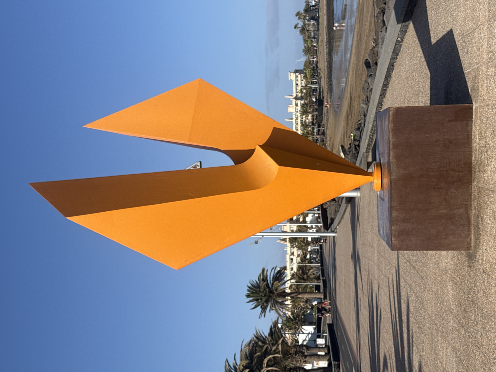 The image shows an orange abstract sculpture located on a paved area next to a beach in Tías, Spain. The sculpture has two triangular shapes curving towards each other and is mounted on a brown rectangular pedestal. Behind the sculpture, there is a view of the beach, buildings, and palm trees. The sky is blue. People are visible on the beach and along the paved area.