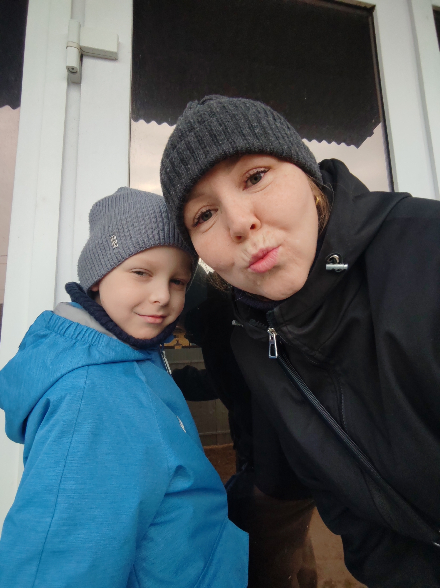 A medium-shot captures a woman and a young boy posing closely for a selfie outdoors. They are standing next to a window with a white frame. The woman, positioned on the right, is wearing a dark gray knitted hat and a black hooded jacket with a zipper. She is slightly angled towards the camera and making a kissing face. The boy, on the left, is wearing a light gray knitted hat and a blue hooded jacket. He is looking at the camera with a slight smile.
The location is determined to be the city of Chaikovsky, Russia. No other information is available.