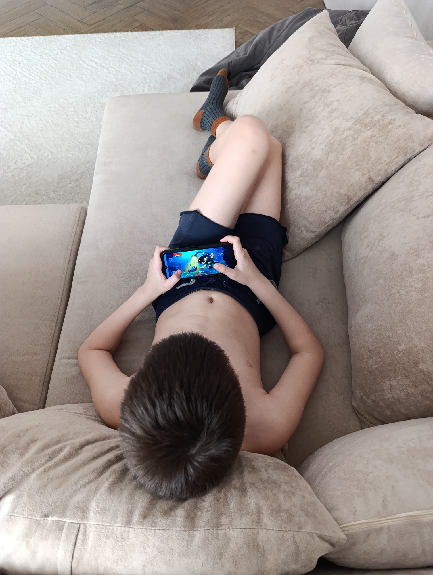 An overhead shot captures a child lying prone on a light beige sectional sofa in an indoor setting, likely a living room in Chaikovsky, Russia. The child, viewed from behind with dark, short hair, is wearing dark blue shorts and gray socks with brown toes and heels. They are engrossed in playing a colorful video game on a black smartphone, holding it horizontally with both hands. The game screen displays cartoonish graphics, with a "100%" indicator visible, possibly for progress or battery life.
The sofa is upholstered in a soft, textured fabric and has several matching pillows, providing a comfortable spot for relaxation. In the background, beyond the sofa, a light-colored, possibly white or off-white, area rug covers a portion of a wooden parquet floor, laid in a herringbone pattern. A dark gray blanket or throw is casually draped over the back of the sofa, near the child's feet. The overall scene suggests a calm and casual moment of leisure. No specific time of day or weather is discernible from the image, but the indoor setting implies a comfortable home environment. The child appears focused and absorbed in the gaming activity.