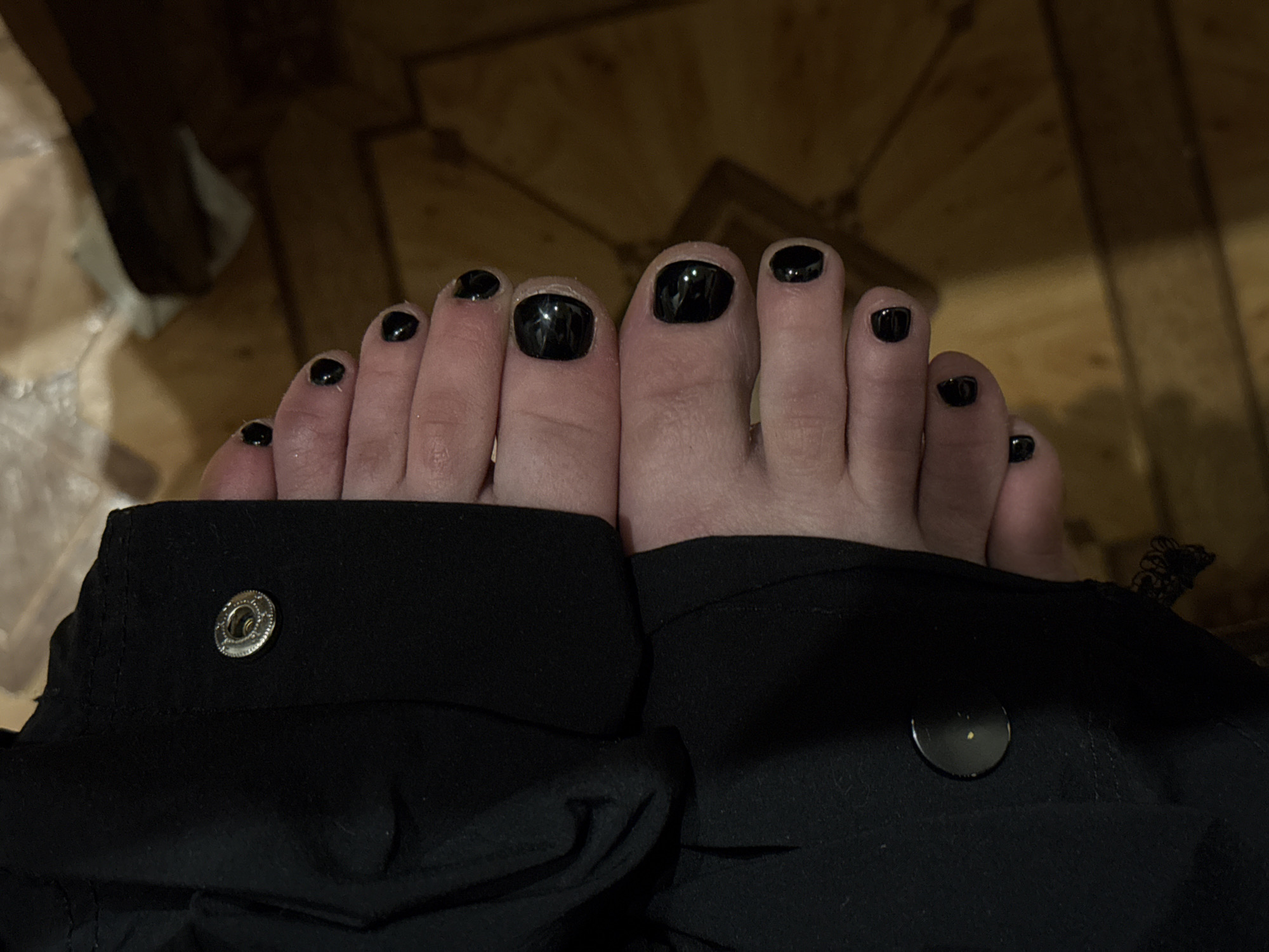 The image depicts a pair of feet with black nail polish on all ten toes. The feet are positioned close together and are partially obscured by what appears to be a black garment, possibly clothing, with a button visible on each side. The background is a wooden surface with a visible pattern. The location is not discernible.