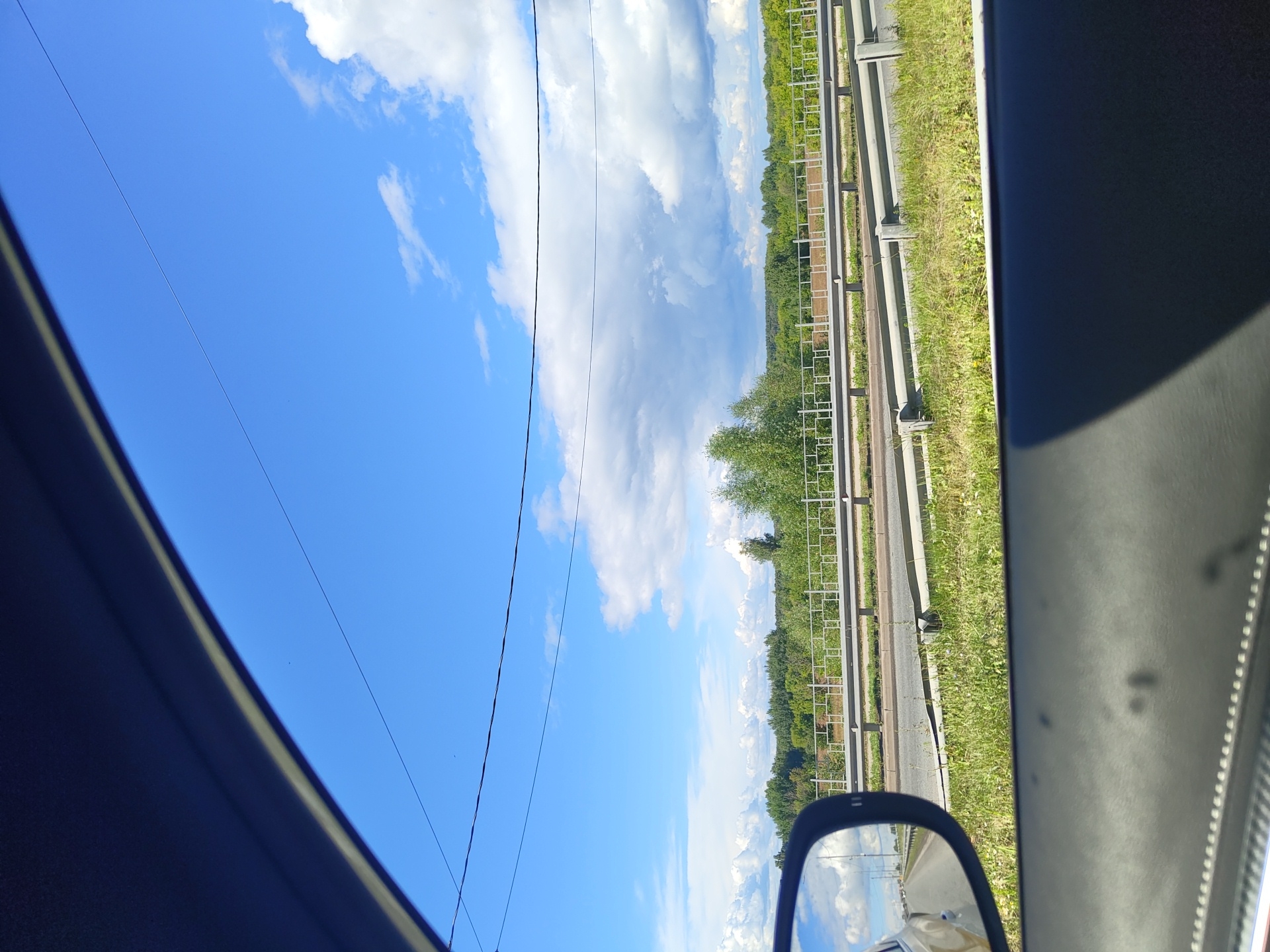 The image is a view from inside a vehicle, looking out the window and a side mirror. The scene appears to be along a highway or main road, with a guardrail separating lanes of traffic. Beyond the guardrail, there is a grassy embankment, followed by a treeline and a vast expanse of blue sky with scattered white clouds.
The side mirror reflects a road, other vehicles, and the sky. The interior of the car is visible in the foreground, including the car's door panel and a portion of the roof. The overall impression is of a sunny day, possibly in a rural or semi-rural area, given the presence of trees and open sky.
It is not possible to determine the exact location of the image within the city of Фруктовая, Russia, based solely on the visual information provided.