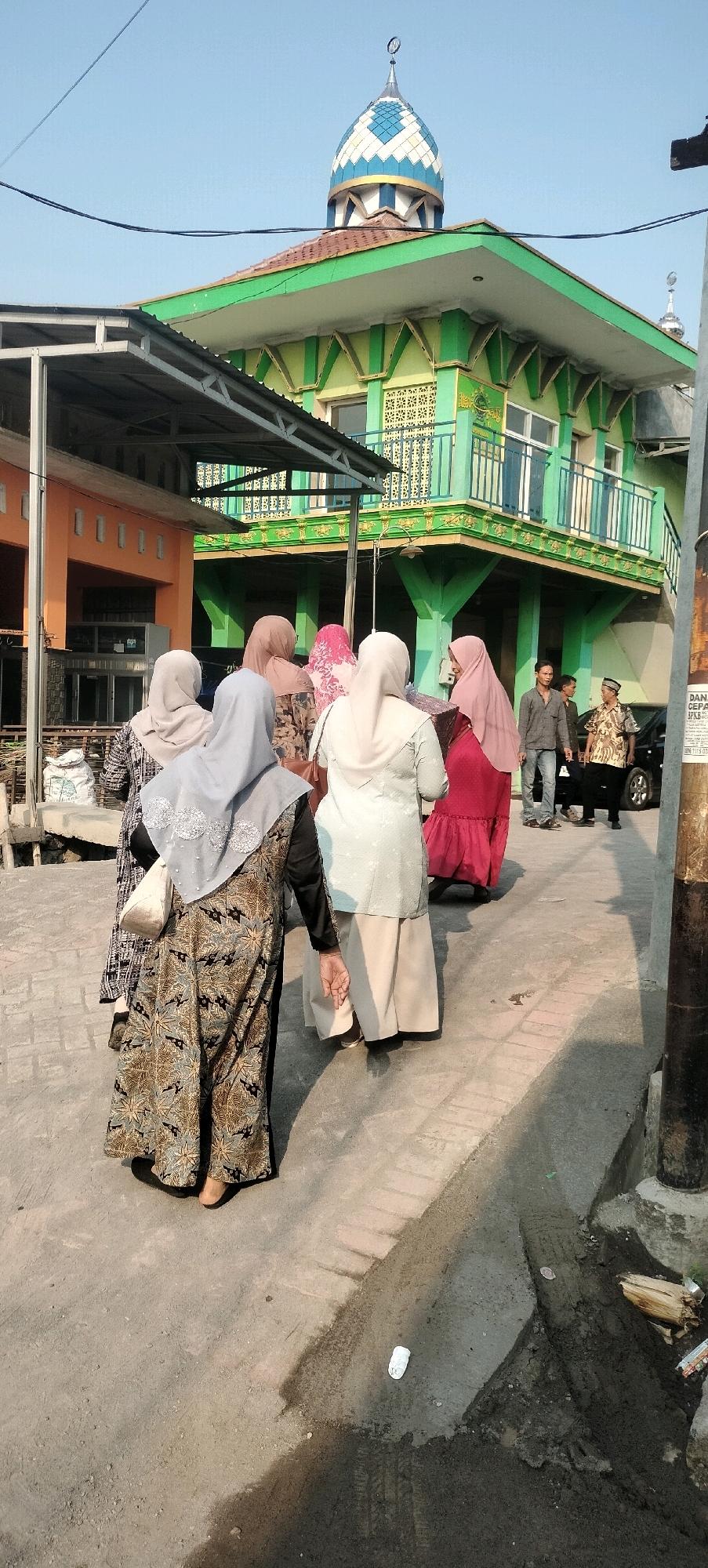 In the city of Semarang, Indonesia, a group of people, primarily women wearing hijabs and long dresses, are walking down a paved road. The women are in the foreground, with their backs mostly to the camera, suggesting they are moving away from the viewer. Their attire is varied in color and pattern, indicating individual choices. Behind them, a vibrant green building with intricate yellow detailing and a distinctive blue and white domed roof stands prominently. This building appears to be a mosque, characterized by its architectural style. The building's facade features windows and decorative elements, contributing to its unique aesthetic. The sky above is a clear, pale blue, suggesting it's daytime. In the background, to the right of the mosque, a dark-colored car is parked, and a few men are standing near it, one of whom is wearing a traditional peci cap. The ground is paved with concrete, and there are some areas of darker, possibly wet or muddy, patches. The overall scene depicts a moment of daily life, possibly after a religious service or a community gathering, in a distinctly Indonesian urban setting.