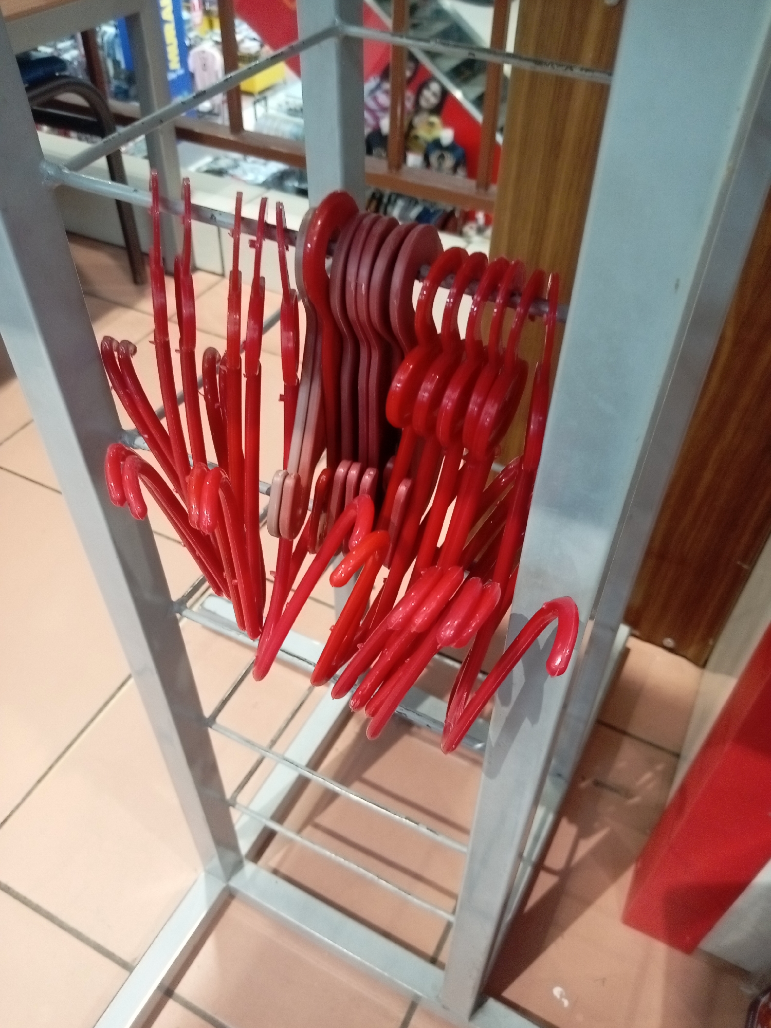 In Batam, Indonesia, a rack of bright red plastic hangers is displayed on a metal frame. The hangers are arranged in several rows, some overlapping. The frame itself is silver and has a grid-like structure at the bottom. In the background, there's a glimpse of a store interior with wooden shelves and clothing items visible. The floor is tiled with large, light brown square tiles.