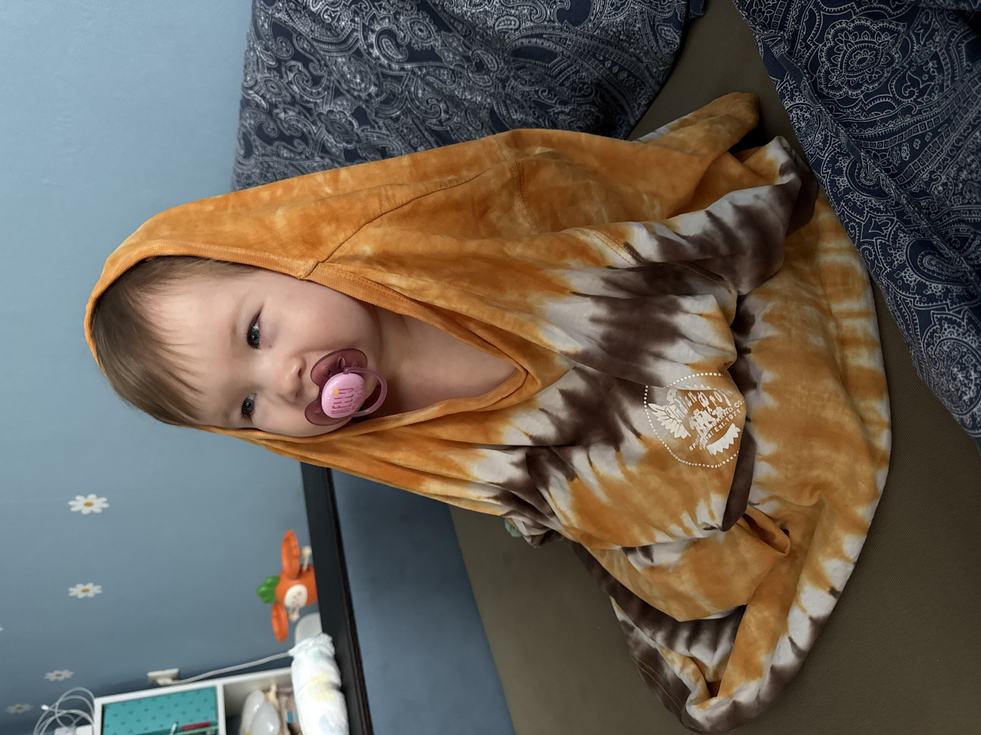 A young child with a pink pacifier is wrapped in a tie-dye hoodie. The hoodie is primarily orange and brown. The child is lying on a surface that appears to be a couch or bed, with a blue patterned textile in the background. The room has blue walls decorated with white flower decals, with some baby supplies are partially visible in the background. It is not possible to determine if this photo was taken in Moscow, Russia based on the available information.