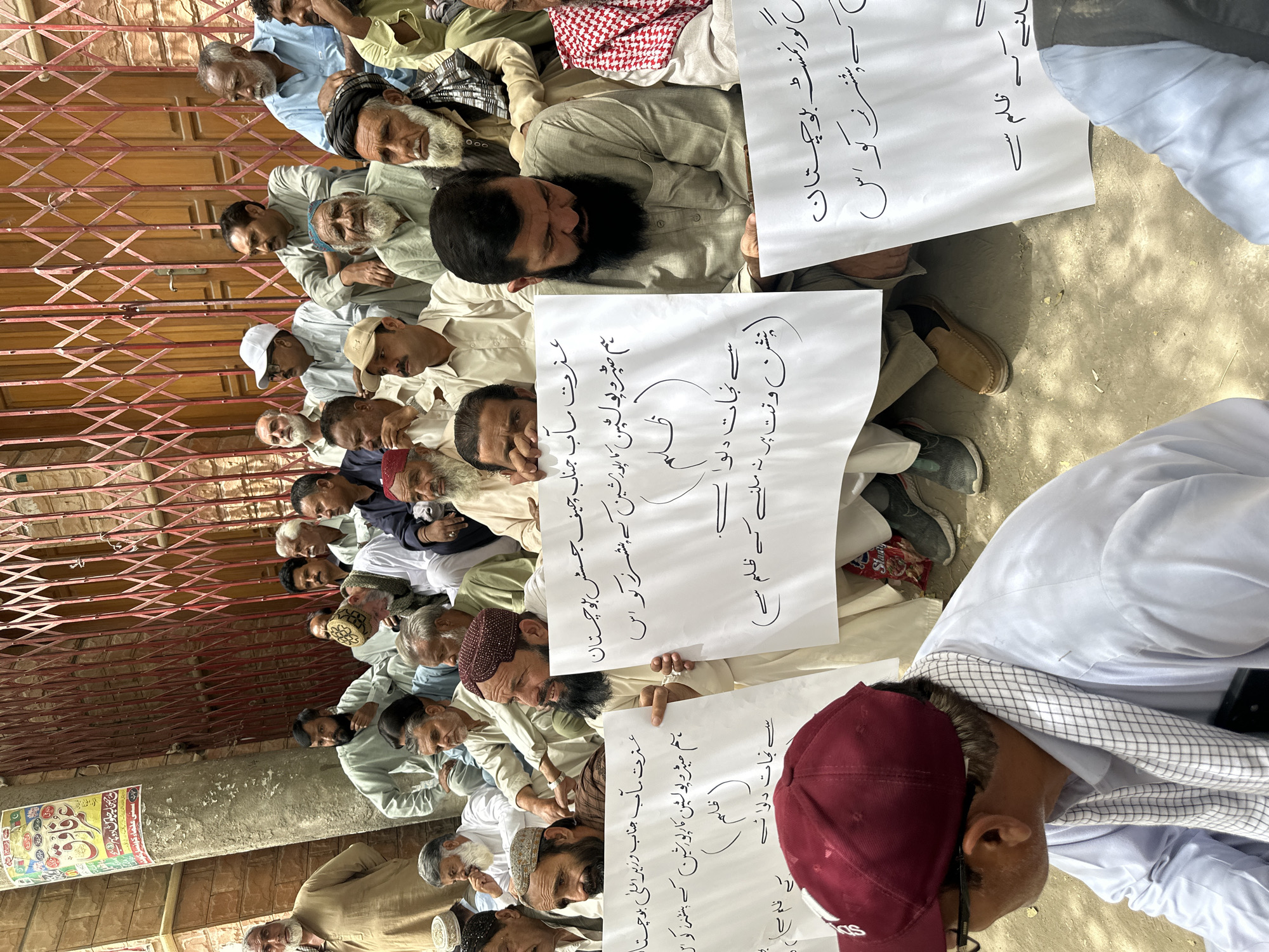 In Quetta City Tehsil, Pakistan, a group of approximately 20 to 25 men are seated on the ground in front of a red metal gate and a brick building. The men are of varying ages and ethnicities, and most are wearing traditional South Asian clothing such as shalwar kameez. Several men are holding up white signs with black Urdu writing. The signs appear to be protest signs. One sign reads "عزت مآب چیف جسٹس بلوچستان" which translates to "Your Honor Chief Justice Balochistan." Another sign reads "ہم ضمبرو پولٹین کے ظلم کے" which translates to "We are from the oppression of Zambrōh Pultain." The men are looking in different directions, some are looking at the camera, some are looking down at their signs, and some are looking at each other. The ground is covered in dirt and gravel. The overall scene suggests a protest or demonstration is taking place.