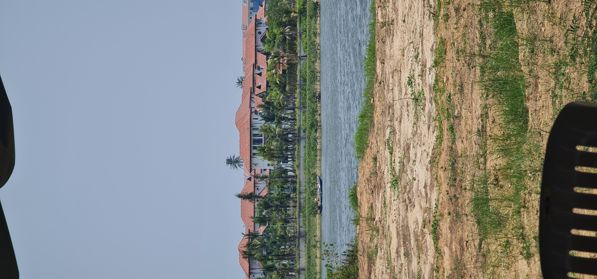 A landscape photograph depicts a view along a waterway, likely a river or channel, in Đà Nẵng, Vietnam. Buildings with orange tile roofs line the opposite bank of the water, interspersed with palm trees. A narrow strip of land vegetated with grass and sparse plants runs along the water's edge on the near side. The sky is light blue. The photo has been rotated 90 degrees, with the sky to the left. Part of a dark-colored object, possibly a chair, is visible in the right foreground.