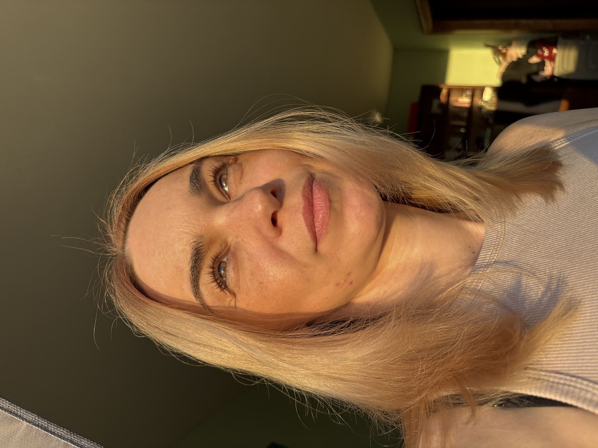 A close-up portrait features a woman with blonde hair and light-colored eyes, tilted slightly on her back. She looks upward and slightly to her right, her expression neutral and contemplative. Strong, warm, golden light, indicative of early morning or late afternoon sunlight, illuminates the right side of her face and hair, creating prominent highlights and shadows. This lighting emphasizes her fair skin, visible pores, and small blemishes on her cheek. Her eyelashes are dark and prominent, and her lips appear naturally pink or with a subtle tint.
She is wearing a light-colored, possibly ribbed, top, visible on her shoulder and upper chest. The background is dimly lit and largely out of focus, suggesting an indoor setting. Hints of furniture or shelves, possibly made of dark wood, and a wall section with a greenish-yellow hue are visible in the blurred background. No visible text is present in the image.
