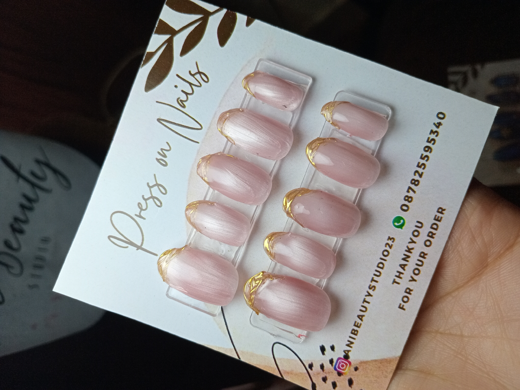 The media file displays a set of light pink, iridescent press-on nails with gold accents, presented on a branded white card. Ten oval-shaped nails are arranged across two transparent plastic holders, five per side. Each nail features a delicate gold outline at its base, creating a subtle reverse French tip effect.
The display card has a cream-colored background adorned with gold foil lettering that reads "Press on Nails" in a cursive script at the top left. Golden leaf motifs decorate the upper right corner. In the bottom right quadrant, black text provides contact and social media details: an Instagram logo followed by "@ANIBEAUTYSTUDIO23," a WhatsApp logo next to the phone number "087825595340," and below it, the phrases "THANKYOU" and "FOR YOUR ORDER." The bottom of the card also features abstract black and brown/gold design elements.
A hand, with visible skin texture, holds the card from the bottom right. The background is dark and out of focus, showing a blurred reflection of another item, potentially another card, with partially visible inverted text such as "Beauty Studio." The image depicts a product presentation, likely for a beauty business, set in the context of North Kuta, Indonesia.