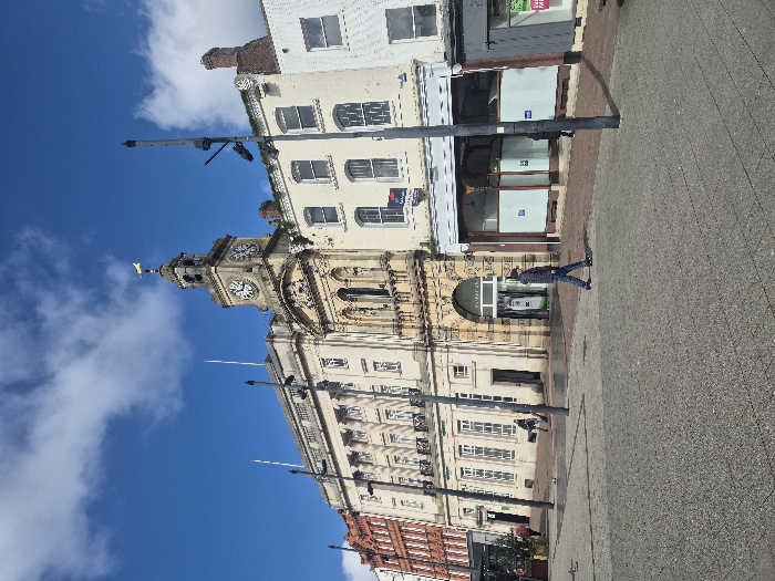 The image displays an outdoor urban scene in Hereford, United Kingdom, specifically depicting the Hereford Town Hall and adjacent buildings, likely viewed from St Owen Street or High Town, under a bright blue sky with scattered white cumulus clouds.
The central and most prominent structure is the multi-story Hereford Town Hall, constructed of light-colored stone. It features a distinguished clock tower topped with a gold weathervane. The clock face indicates a time of approximately 12:45. The building exhibits ornate architectural details including arched entrances, classical columns, statues within niches, and multiple windows. To the immediate left of the Town Hall is a section of Hereford Shirehall (now Council Offices), a similarly light-colored building with a classical facade, numerous windows, and a balustraded upper section. Further left, a multi-story building constructed of orange-red brick is partially visible.
To the right of the Town Hall, a sequence of multi-story buildings extends. The first building, directly adjoining the Town Hall, is light-colored and features several windows; an upper window displays a "TO LET" sign. Its ground floor presents a recessed, ornate storefront with large windows covered by white paper and blue signs. To the far right, another white-painted building with a grey ground-floor retail unit is partially observed.
The foreground and midground consist of a paved public area with sections of grey and reddish-brown paving stones. Several tall, black, multi-armed streetlights are interspersed throughout the scene, casting discernible shadows to the right.
Approximately six human figures are present. In the mid-foreground, a male individual wearing a dark jacket and jeans walks from left to right across the paved area. Three other indistinct figures are visible walking in the mid-left background near the Shirehall building. A single figure is partially obscured by a streetlight pole on the right side. Additionally, several pigeons are scattered on the paved ground in the mid-left. No direct interactions between individuals are evident.