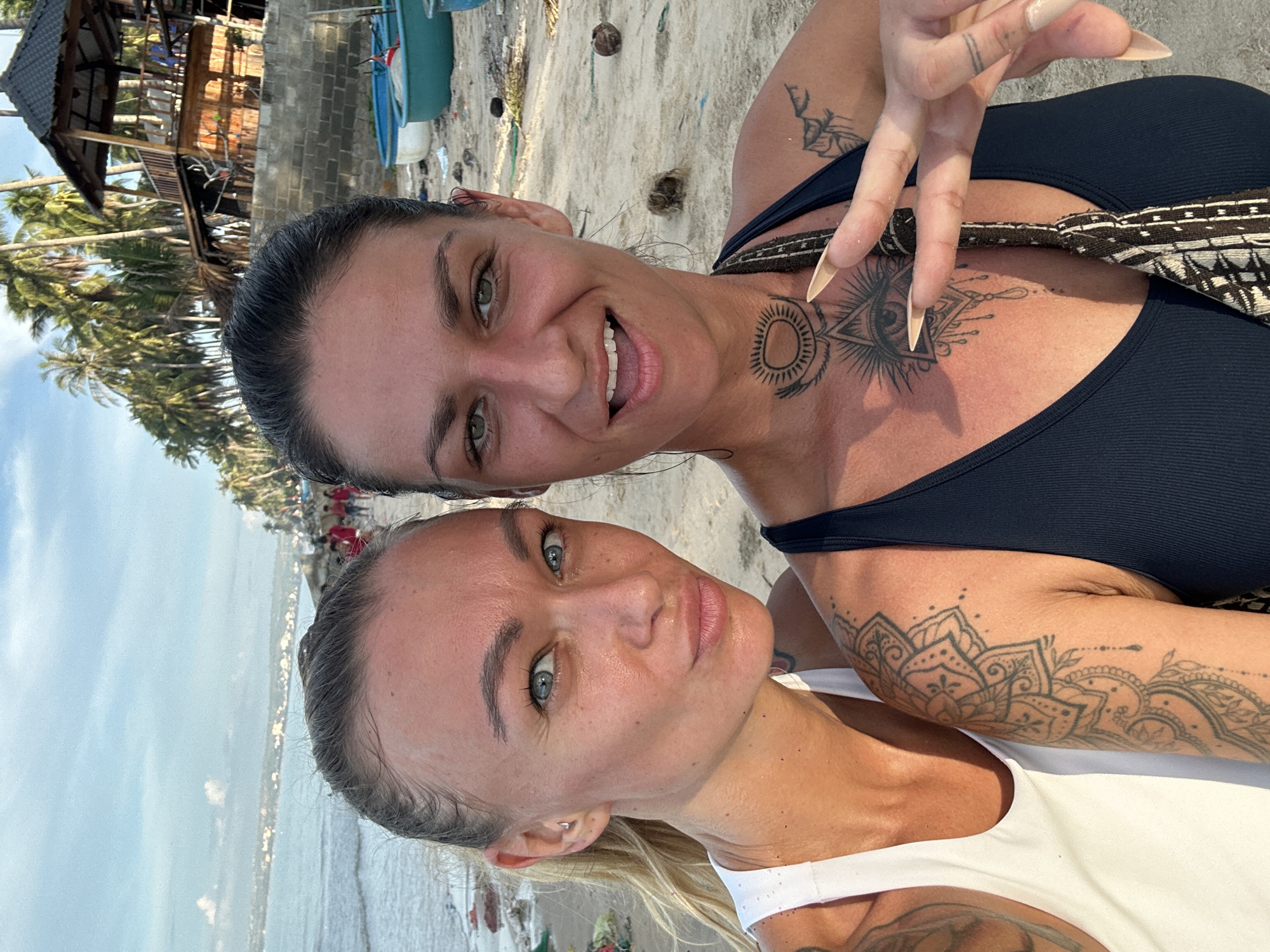 Two women are posing for a selfie on a beach in Mũi Né, Vietnam. The woman on the left has blonde hair pulled back in a ponytail and is wearing a white tank top. She has blue eyes and is looking towards the camera with a slight smile. The woman on the right has dark hair pulled back and is wearing a dark tank top. She has green eyes and is smiling broadly at the camera, holding up two fingers in a peace sign. Both women have tattoos on their arms and chests. The background shows the ocean with small waves, a sandy beach with scattered debris, palm trees, and a wooden structure. The sky is partly cloudy.