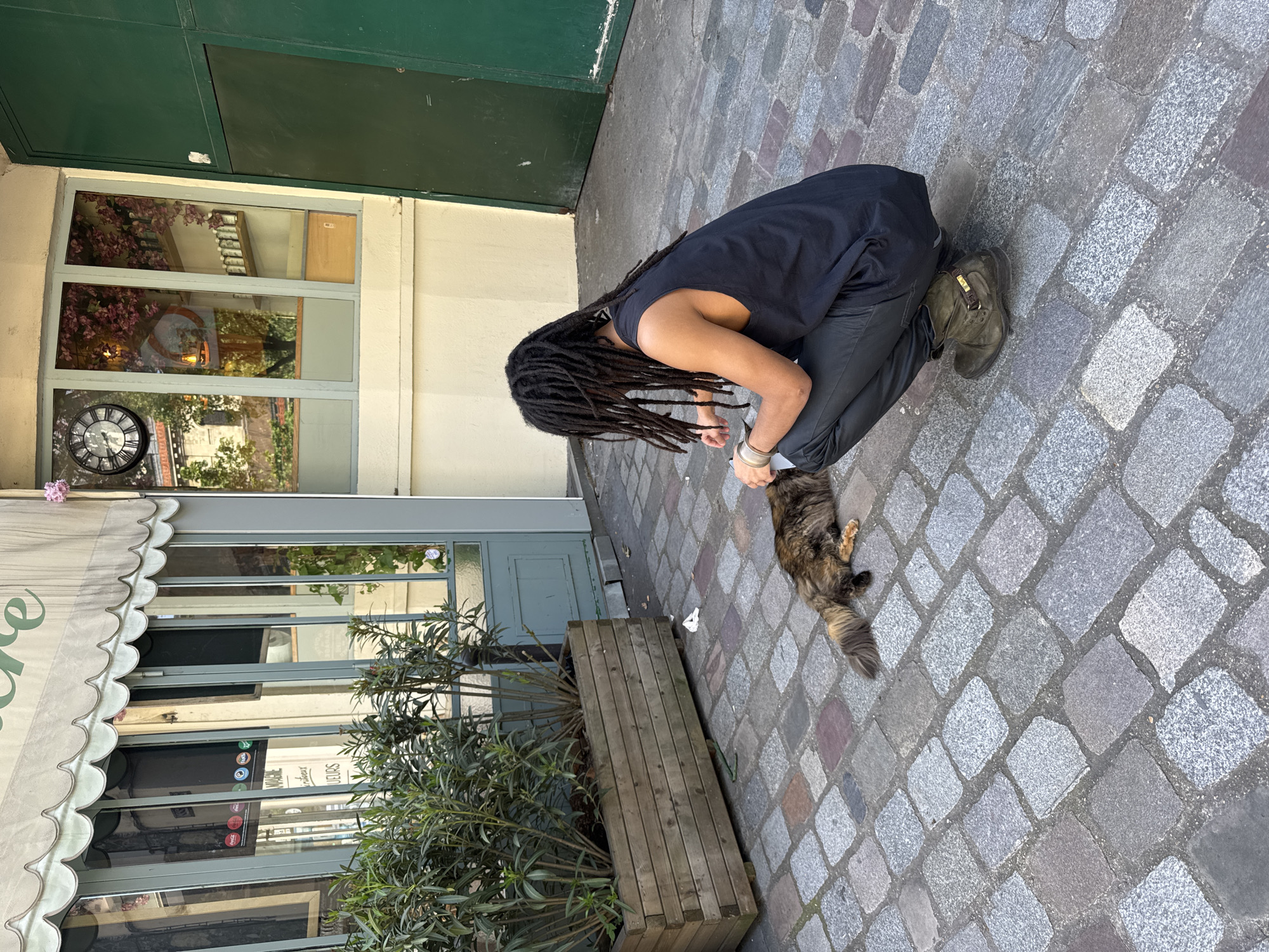 A person with dreadlocks wearing a black tank top and dark pants kneels on a cobblestone street, interacting with a long-haired brown and black cat. The person appears to be offering something to the cat, possibly food. The scene is framed by the facade of a building with a green door, a window reflecting a clock and interior elements, and a striped awning over a shop window displaying stickers. A planter filled with green plants sits nearby on the sidewalk. The location appears to be in Paris, France, suggested by the architecture and style of the storefront. The time is afternoon.
